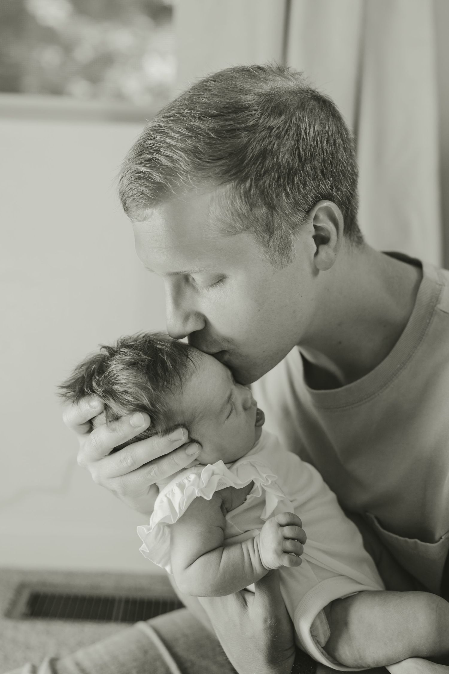 Black and white photo of father kissing newborn baby girl on forehead with tender affection.