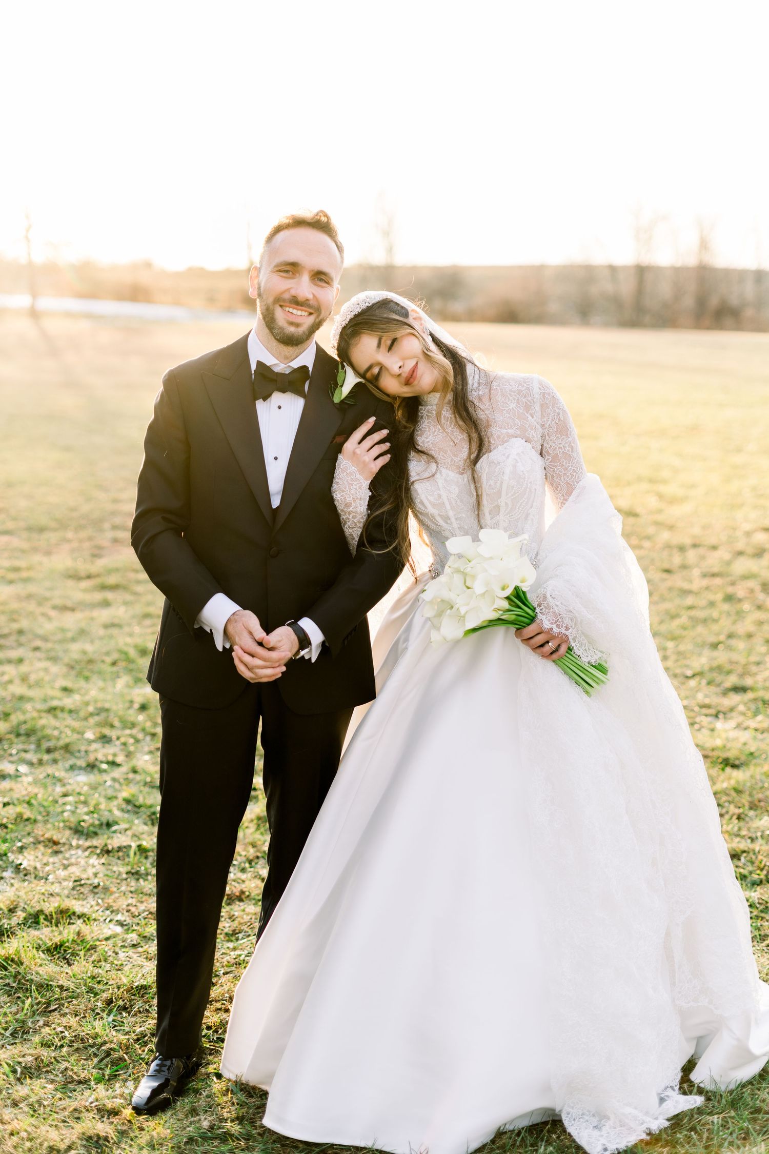 Smiling bride and groom pose together at golden hour outdoors, bride in lace gown holding white calla lily bouquet.