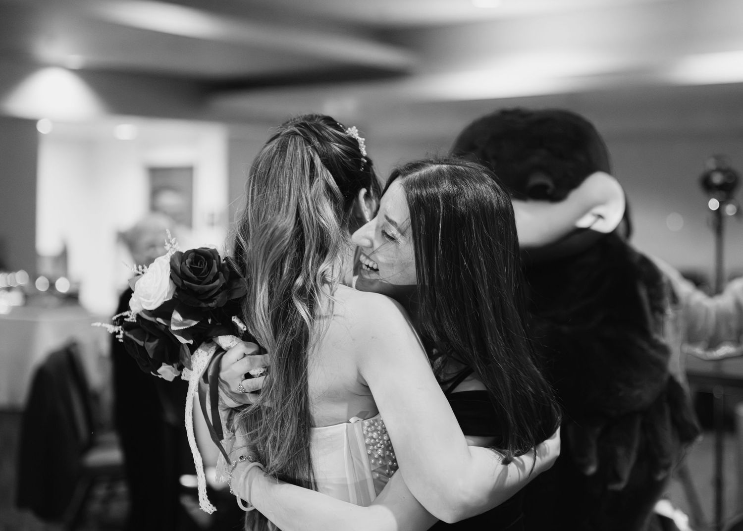 Bride embracing a guest while holding a bouquet at a wedding reception in black and white.