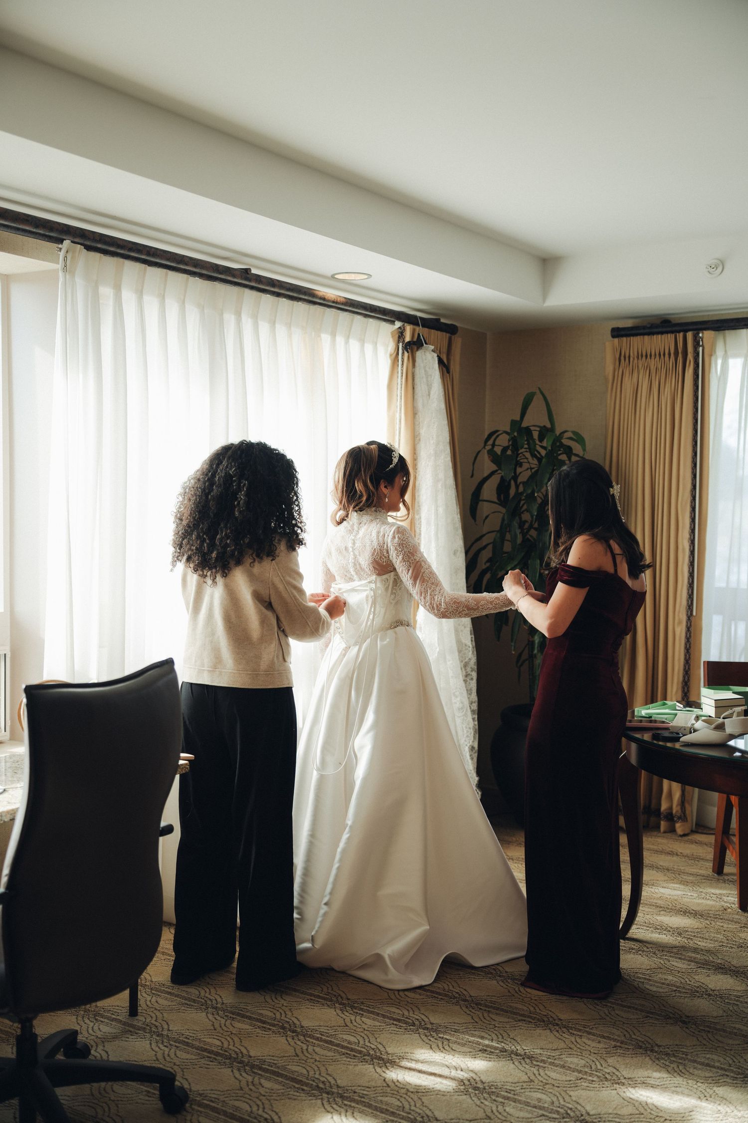 Two bridesmaids help a bride in a lace wedding gown get dressed near a bright hotel room window.
