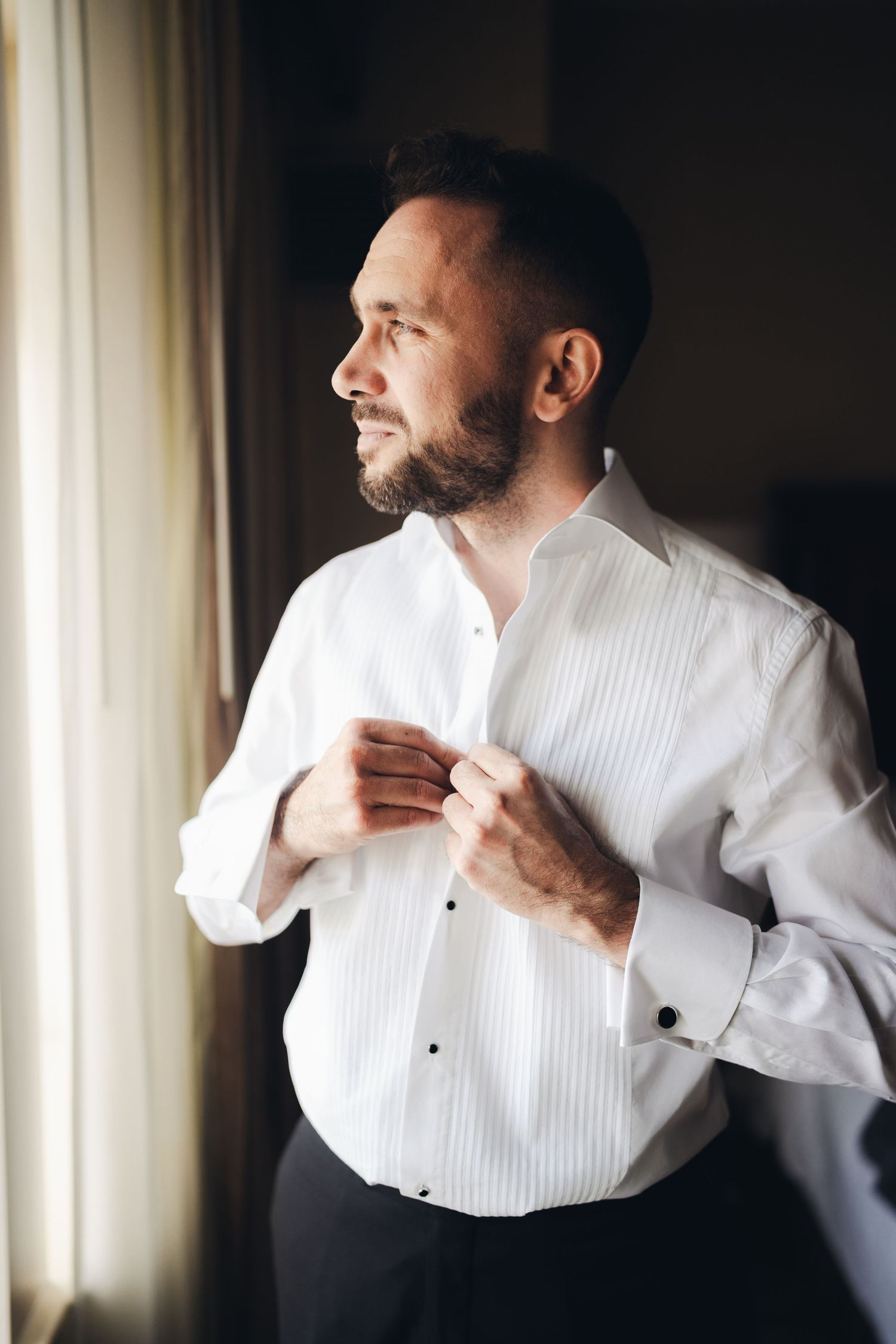 Handsome bearded groom in white tuxedo shirt buttoning cufflinks while gazing out window before wedding.