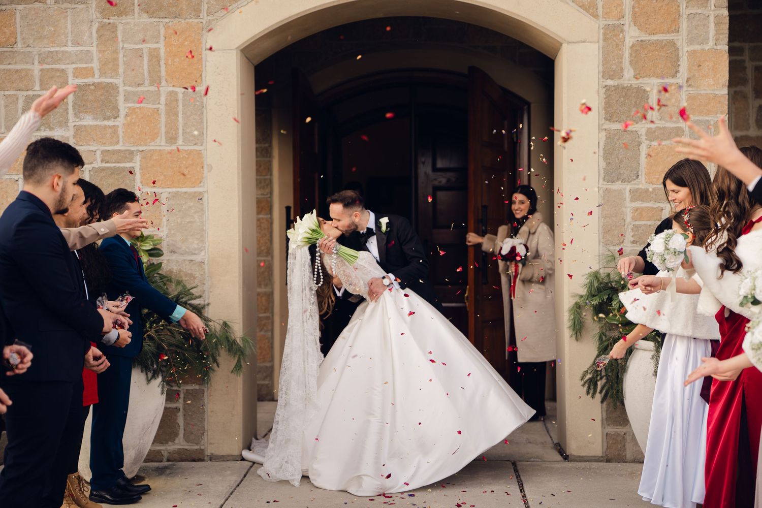 Newlyweds kissing as guests toss flower petals outside stone church.