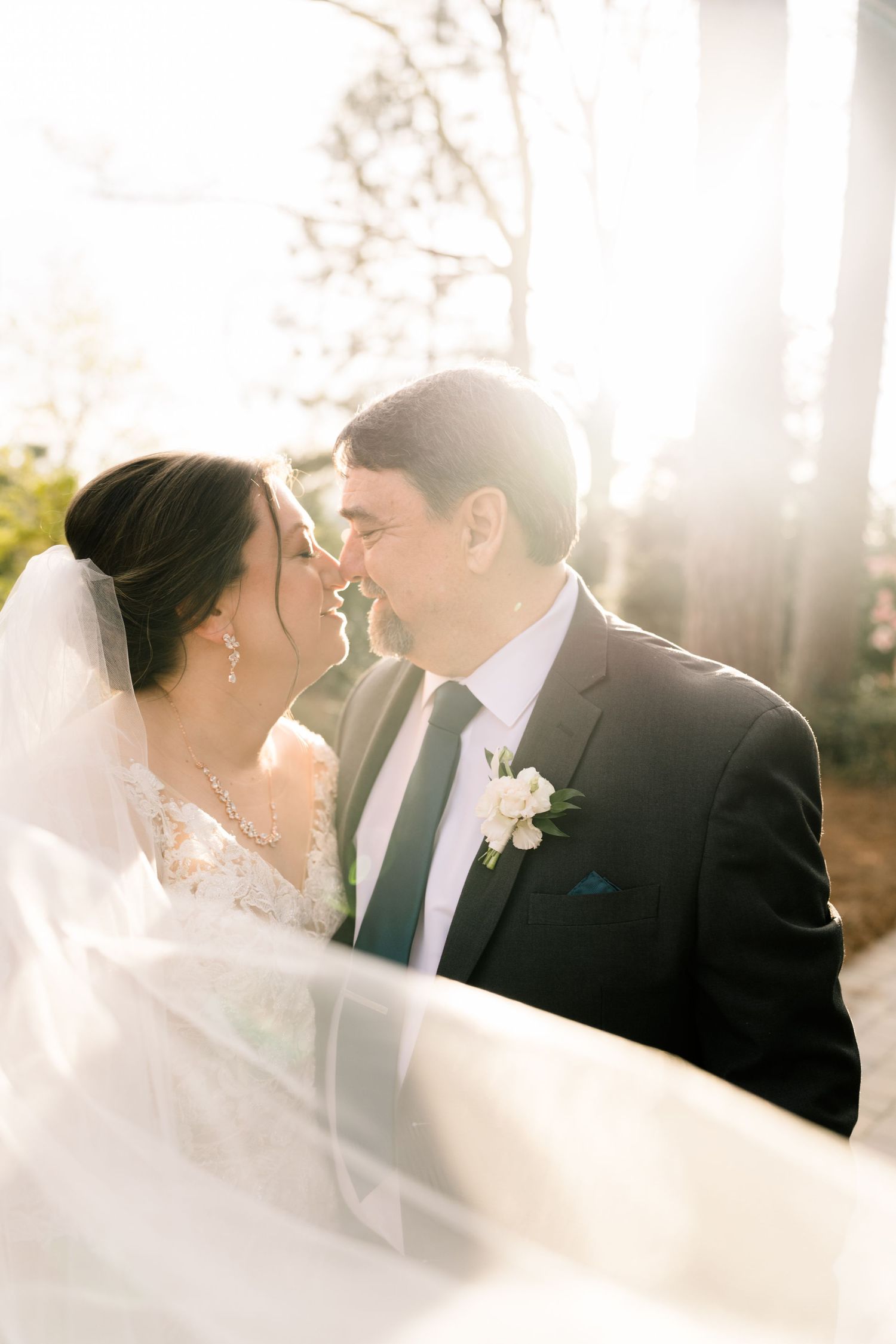 Bride and groom sharing a tender almost-kiss with flowing veil and radiant backlit sunlight.