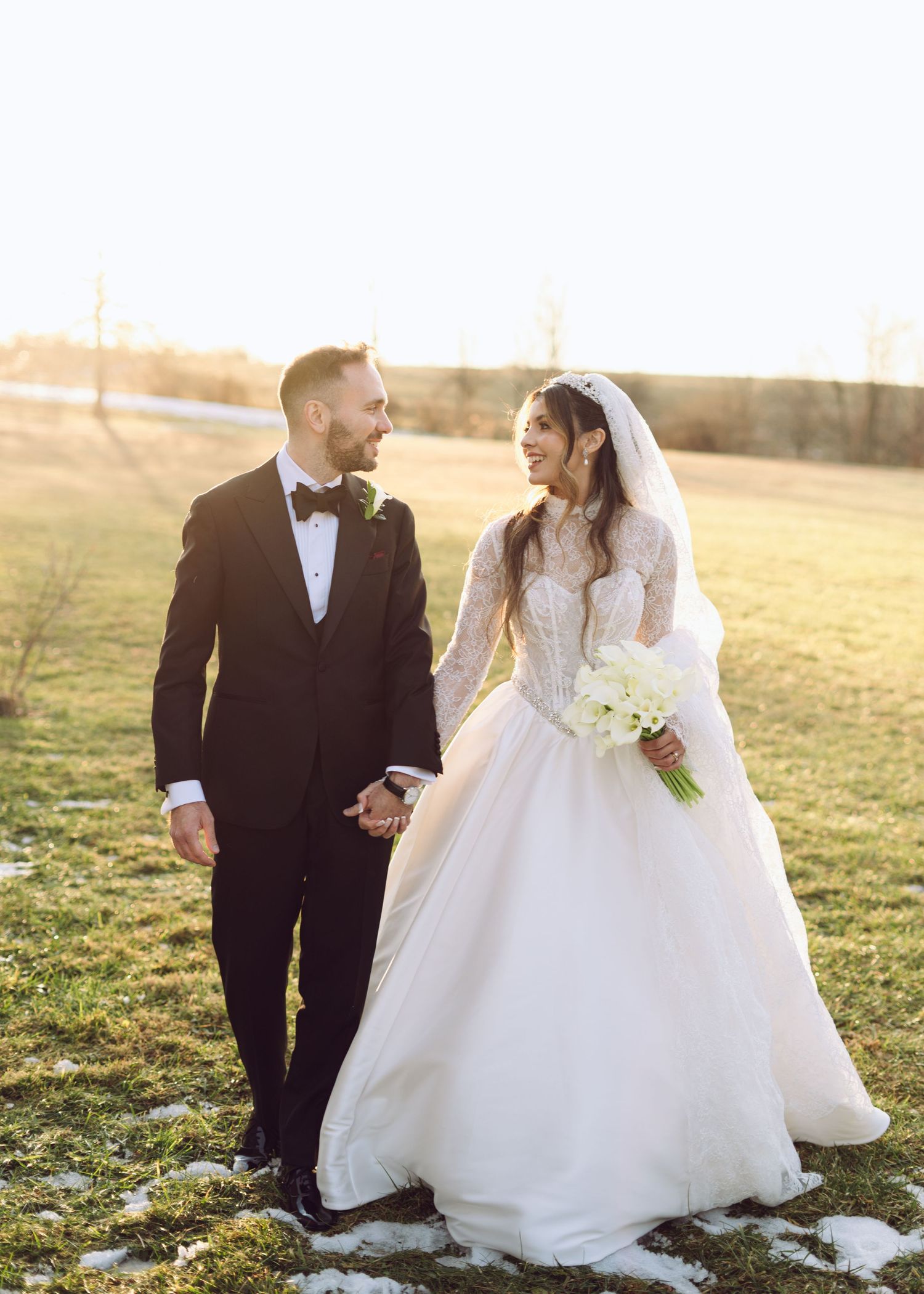 Joyful bride and groom walking hand in hand in a sunlit field, bride carrying white calla lilies.
