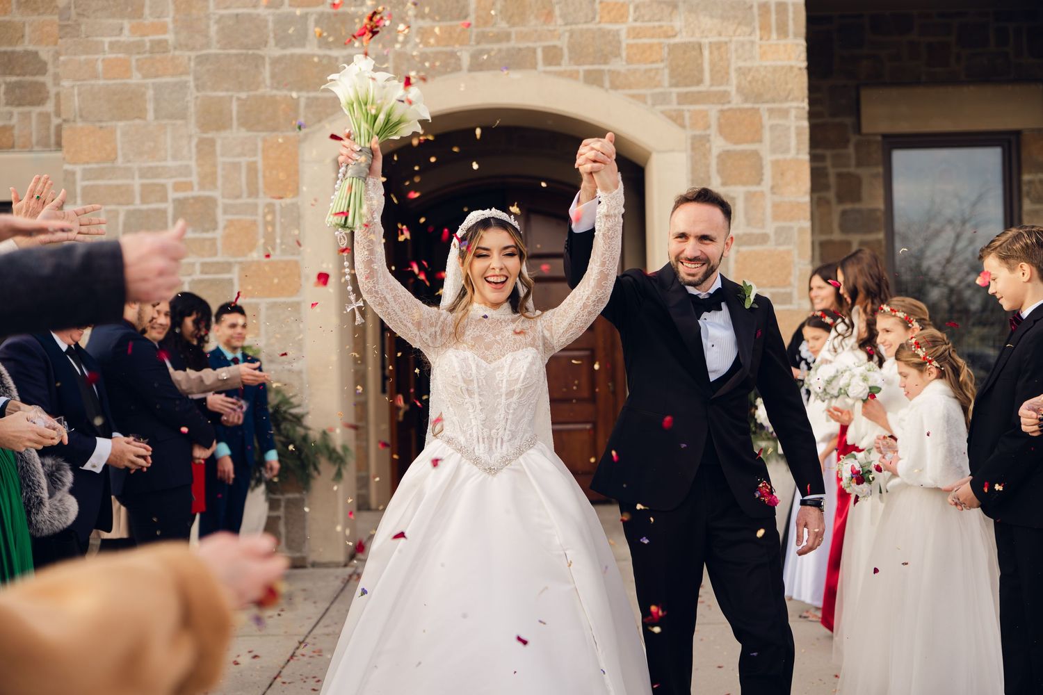 Joyful newlyweds exit church as guests shower them with red flower petals in celebration.