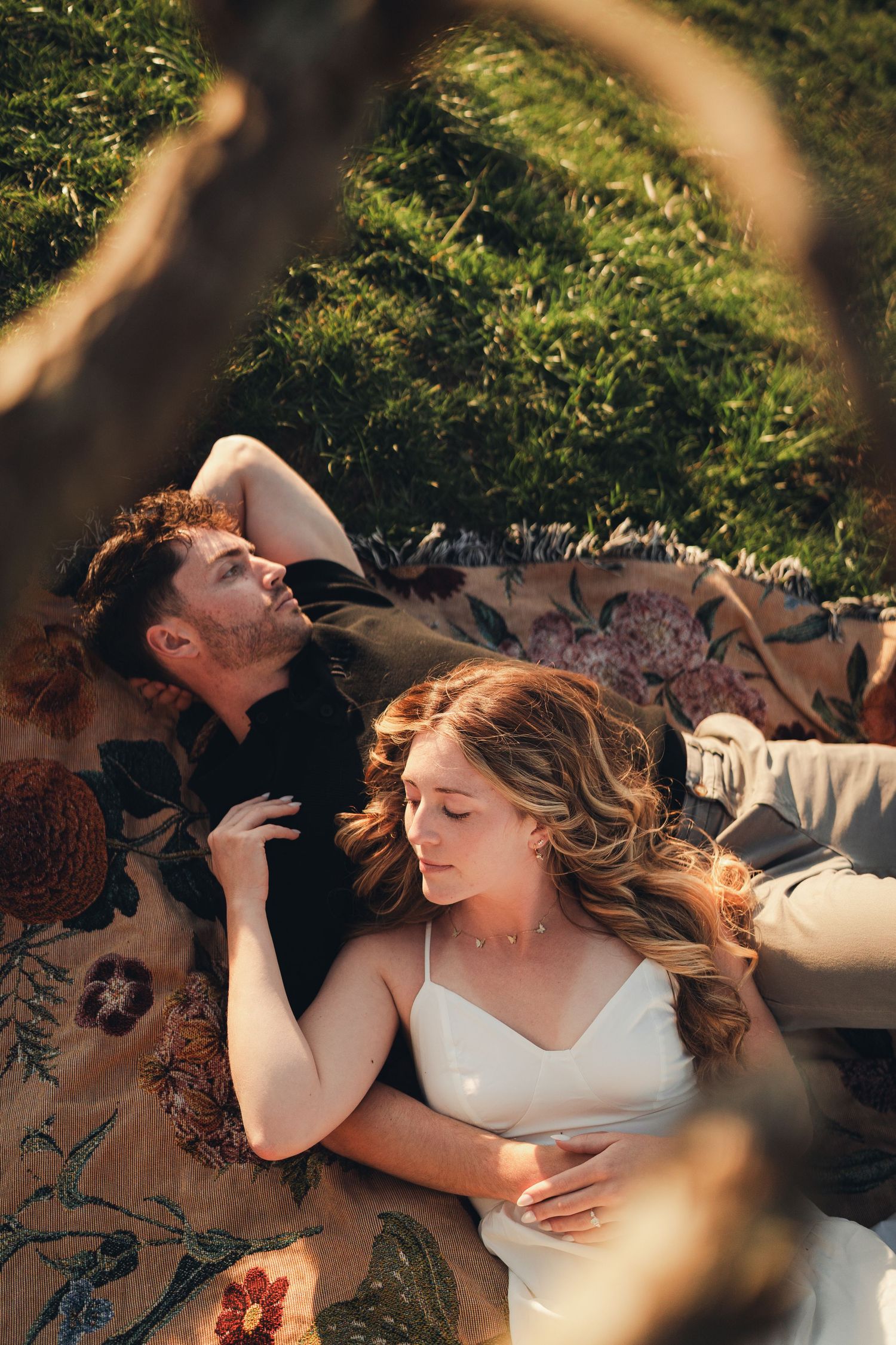 Bride and groom lying on a floral blanket on a grassy hillside in warm sunlight.