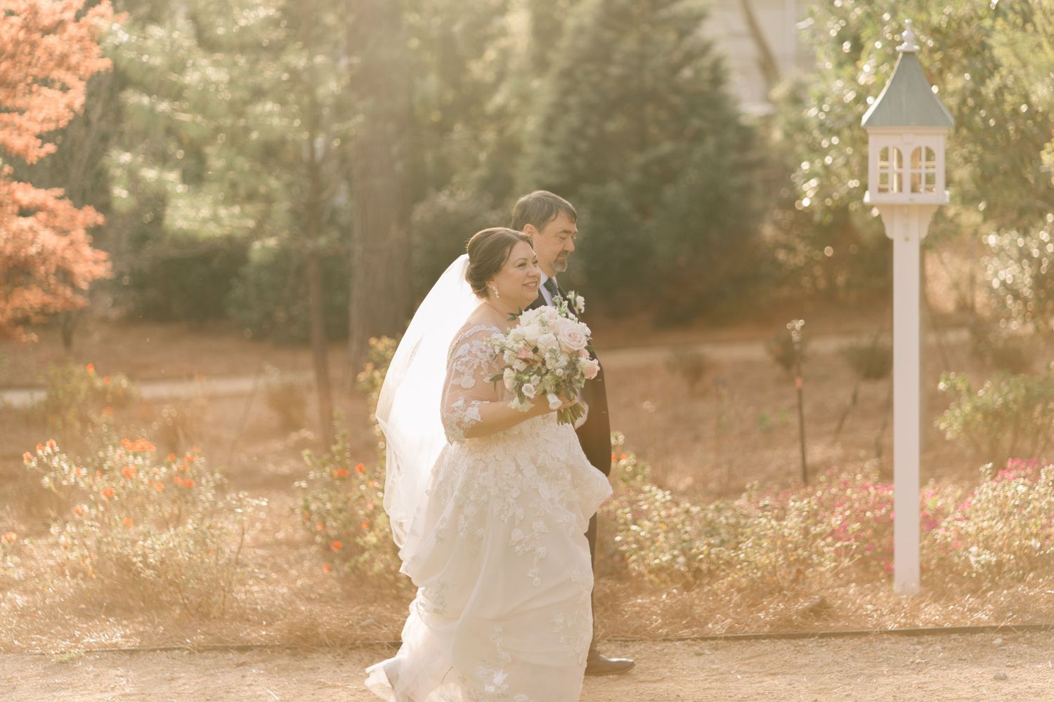 Bride and groom walking hand in hand through a sunlit garden path with autumn foliage.