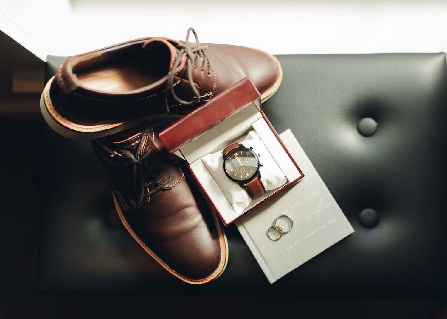 Brown leather dress shoes, a classic wristwatch in a box, and gold wedding rings arranged on a black leather surface.