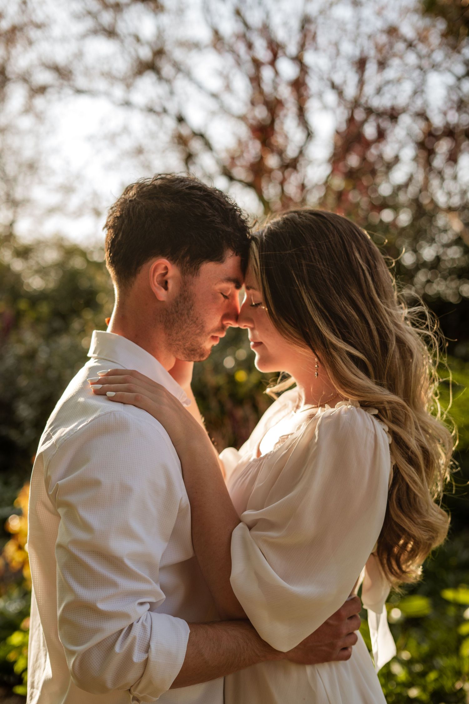 Couple embracing with foreheads touching in a warm sunlit garden, sharing an intimate romantic moment.
