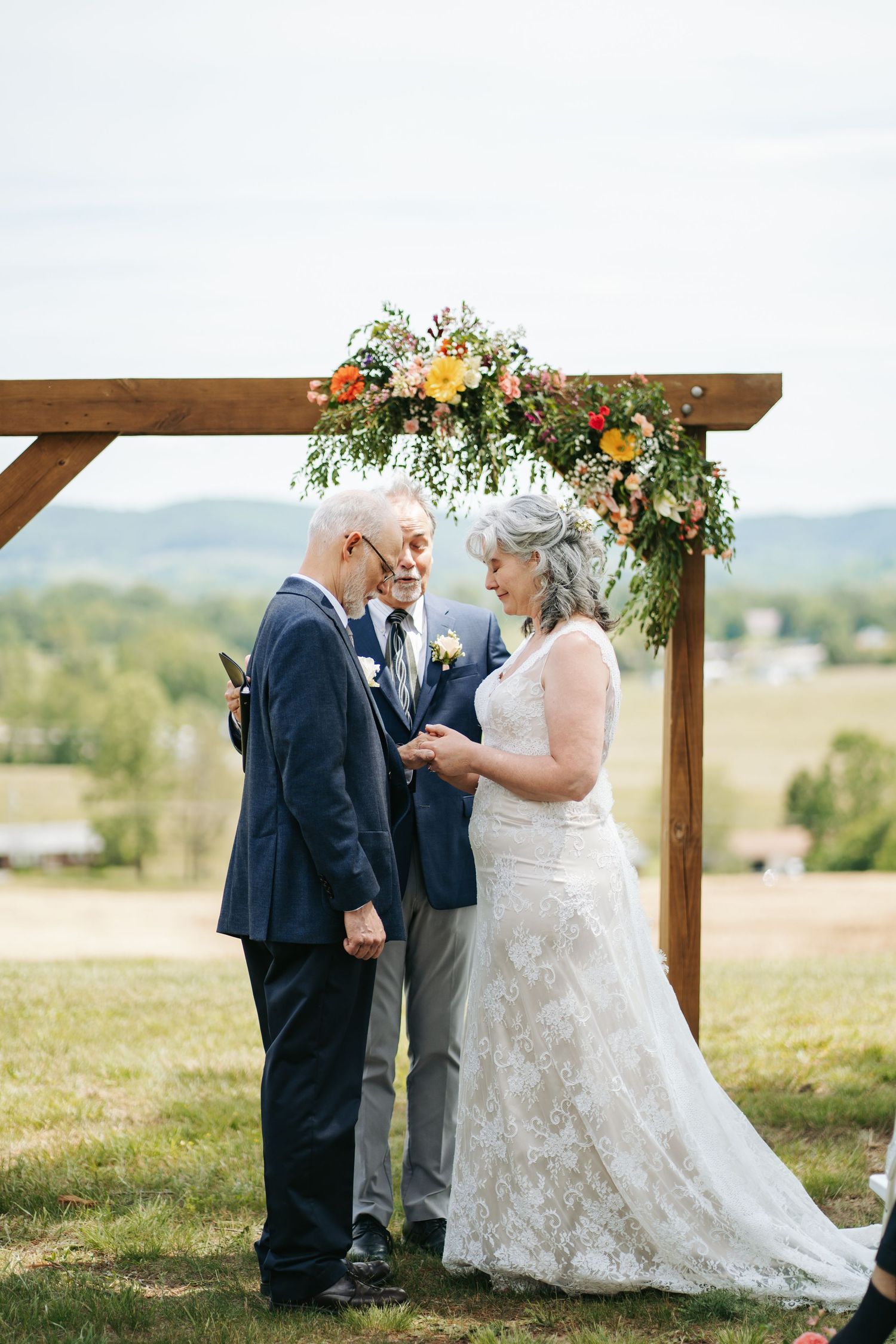 Older couple exchanging vows under floral wooden arch with scenic mountain valley backdrop.