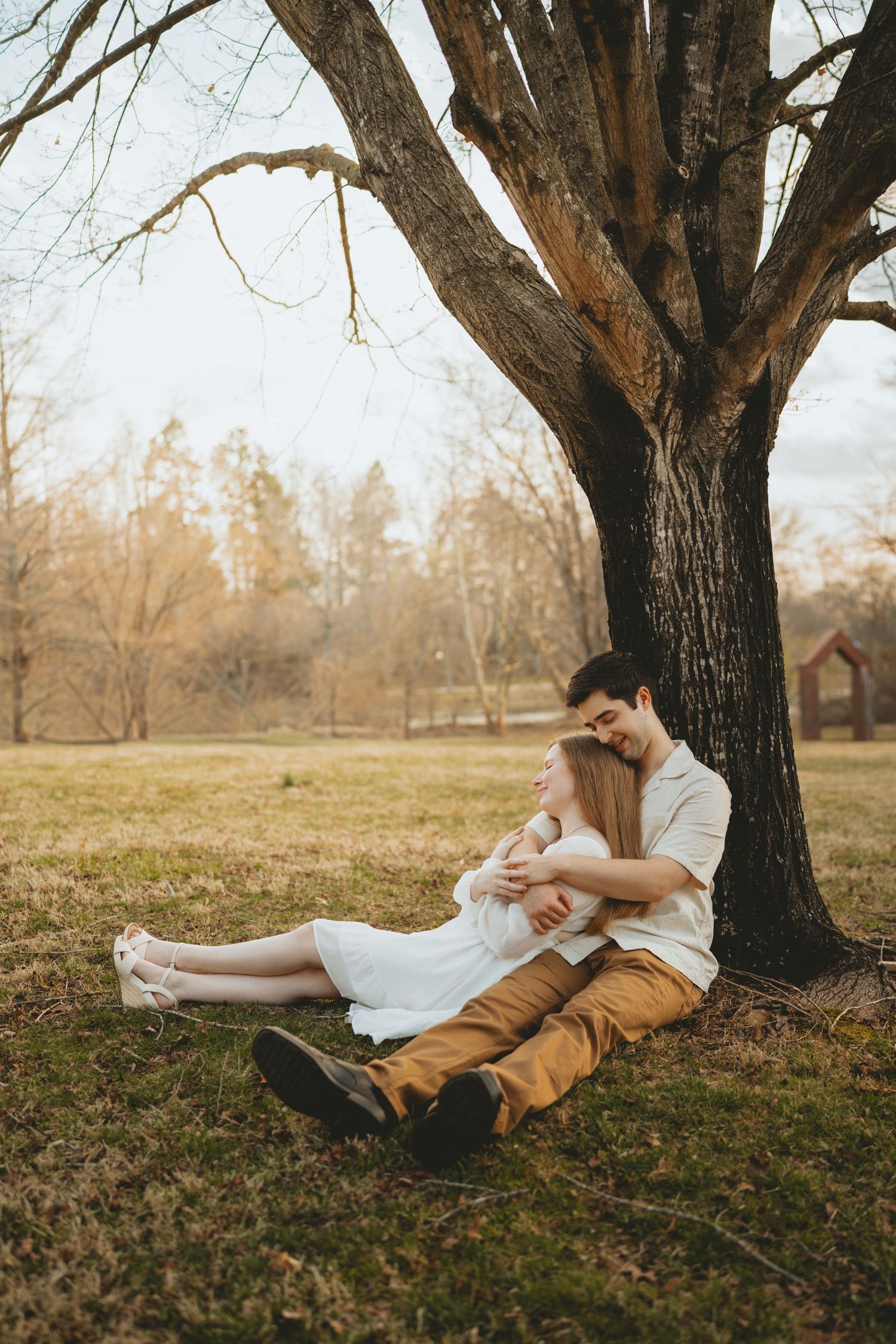 A couple sits cuddled together against a large tree trunk in a serene autumn park landscape.