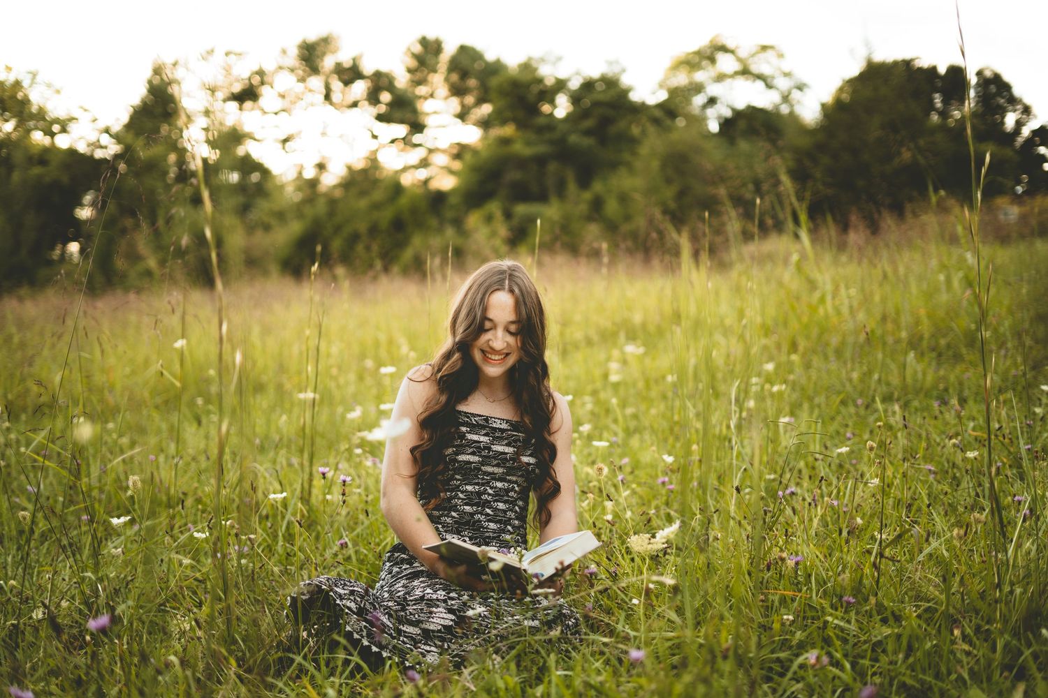 Young woman with curly hair reading book while sitting in lush green meadow.