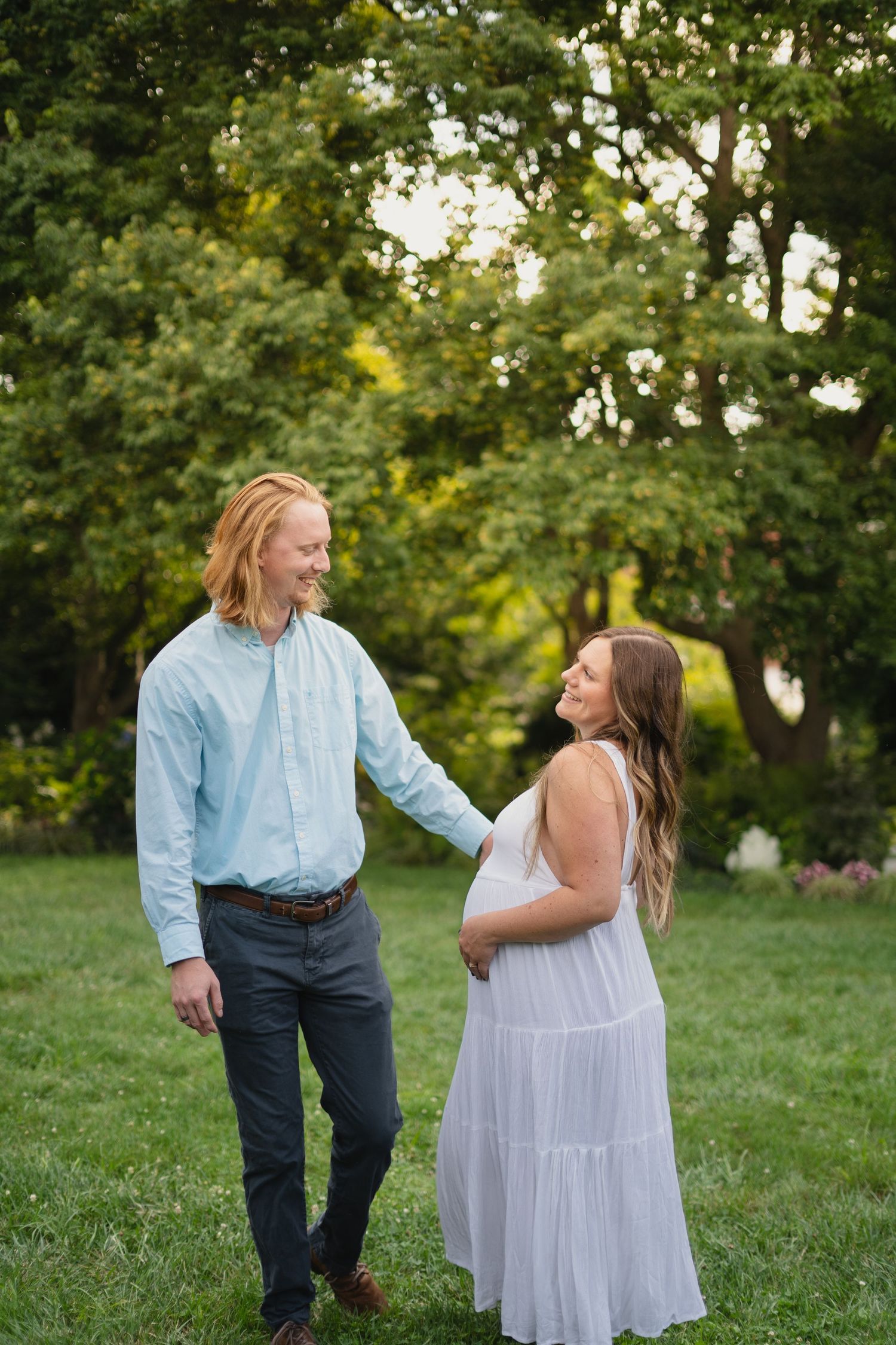 Smiling couple holding hands in a lush green park, pregnant woman in white dress and man in light blue shirt.