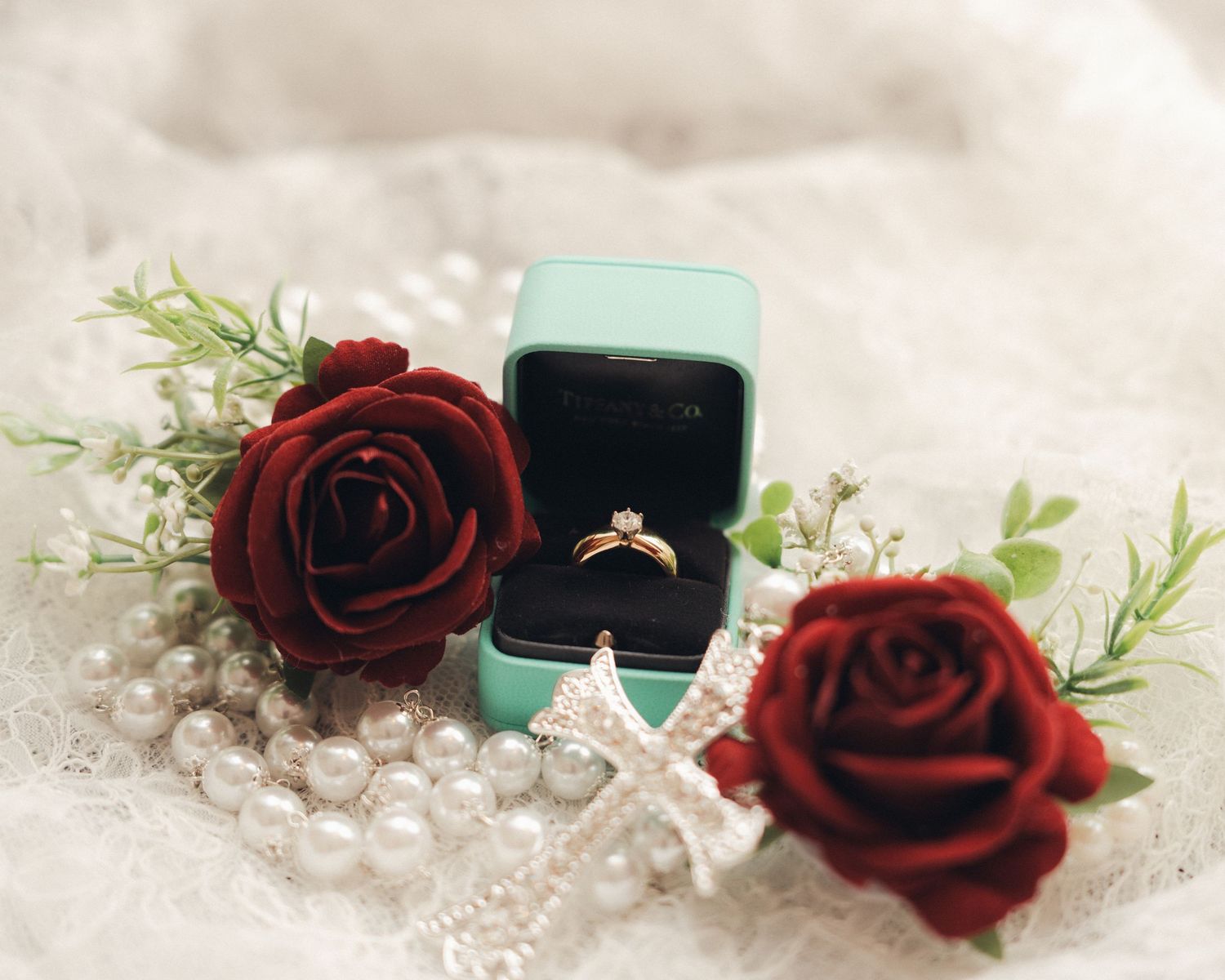Gold engagement ring in teal box surrounded by red roses and pearl necklace on lace fabric.