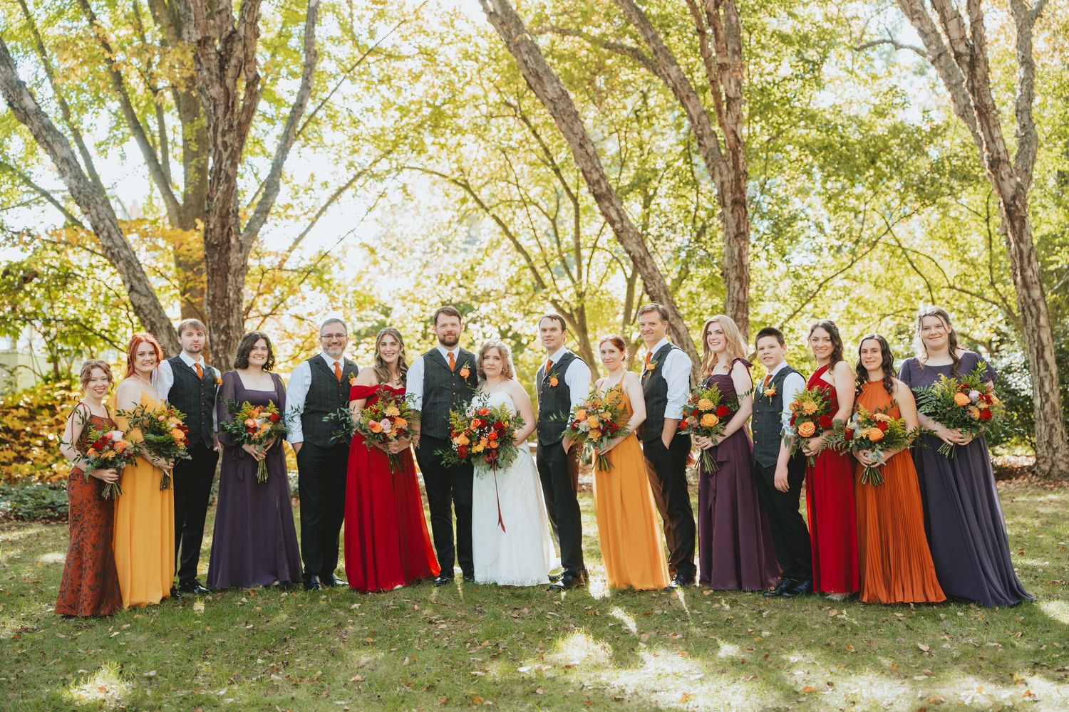 Large wedding party in autumn-themed mismatched bridesmaid dresses of red, purple, and orange standing outdoors.