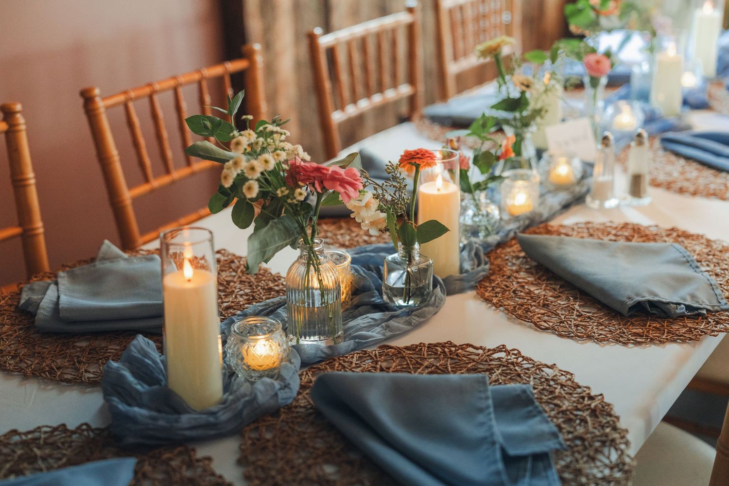 Elegant wedding reception table with blue napkins, candles, floral centerpieces, and a brown sequin runner on white linen.