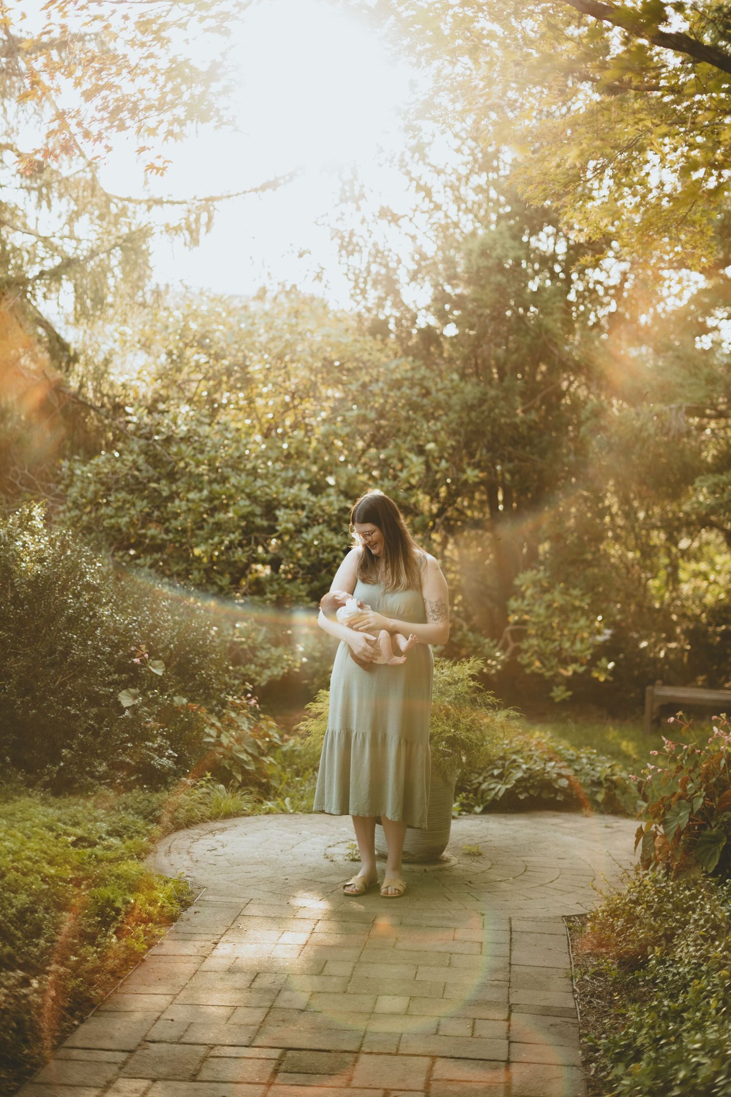 A mother in a sage green dress cradles her newborn baby while standing on a sunlit garden pathway.