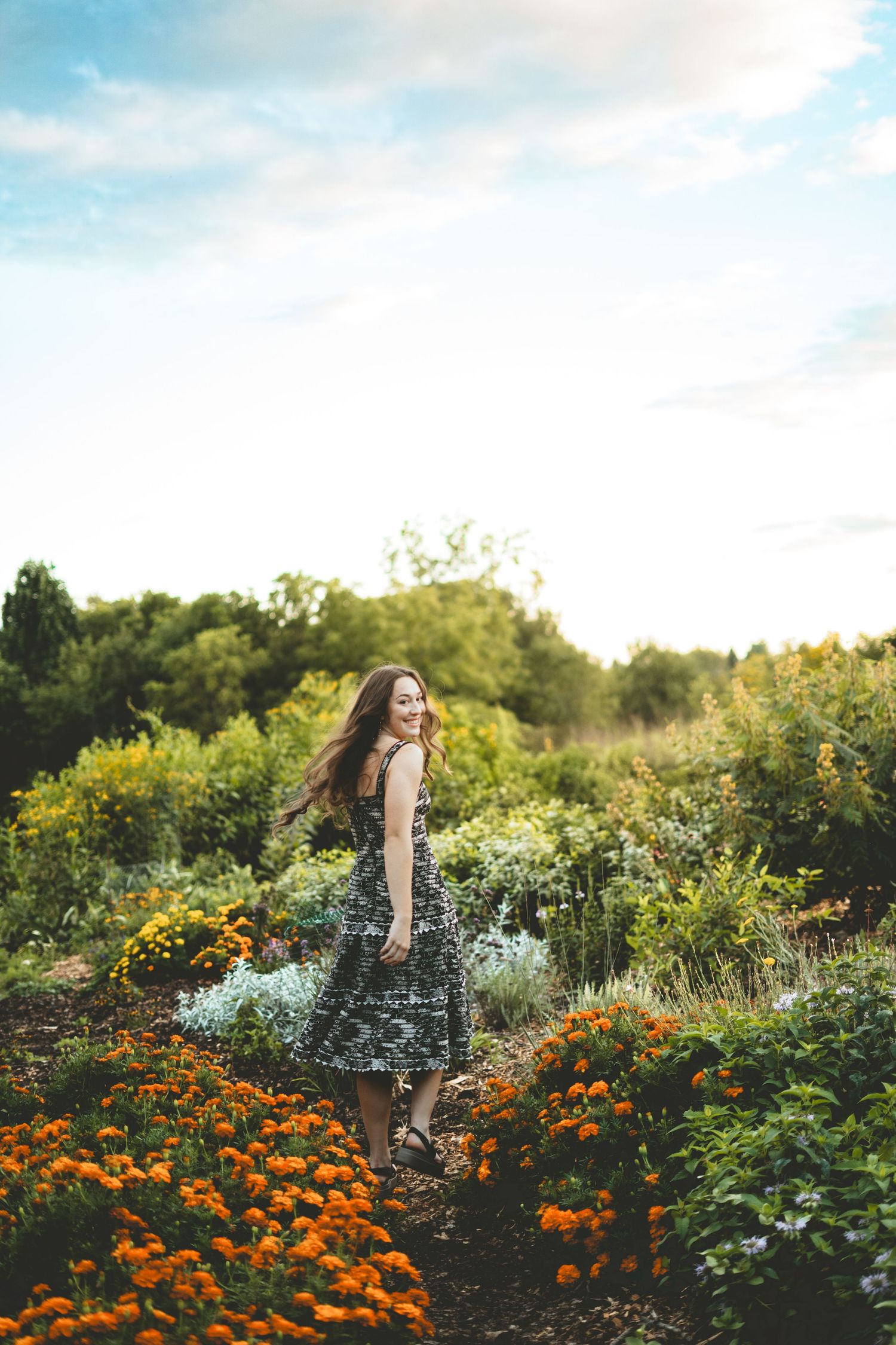 Young woman in floral dress walks through vibrant orange marigold garden at sunset.