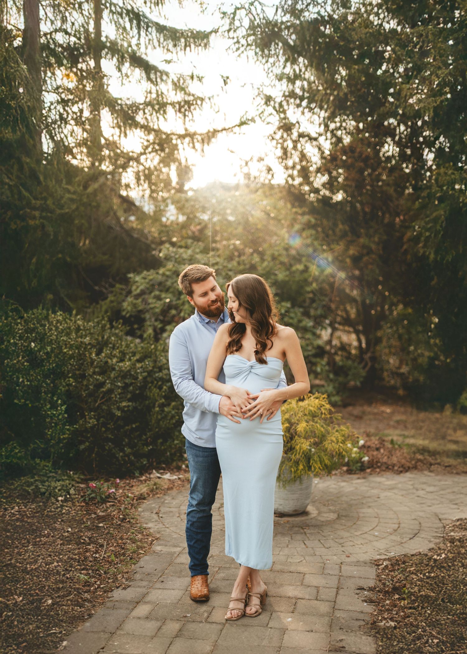 Couple posing together on a garden path, hands on baby bump, in a lush green outdoor maternity portrait.