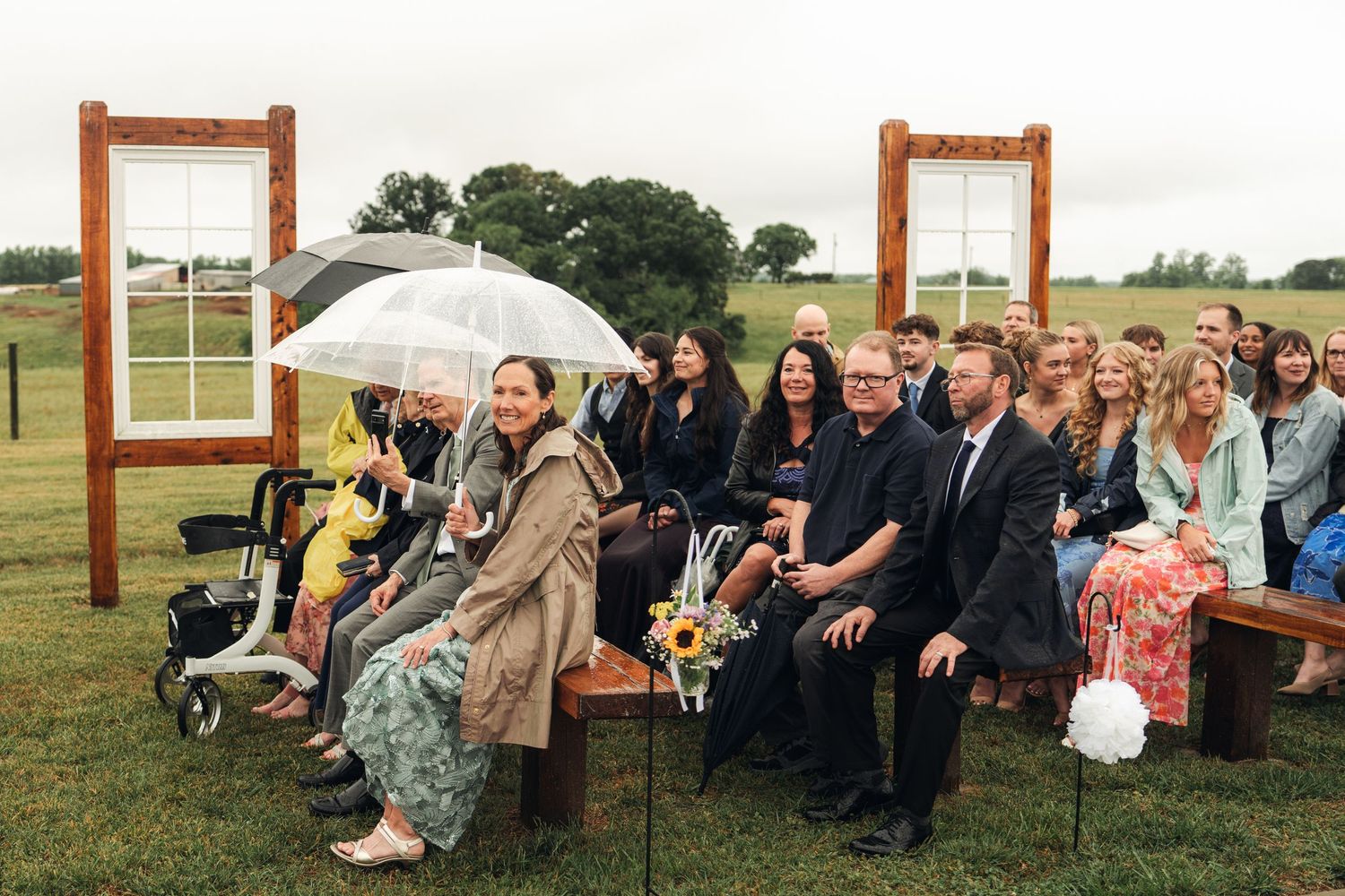 Wedding guests seated on wooden benches outdoors in a field, some holding umbrellas on a rainy day.