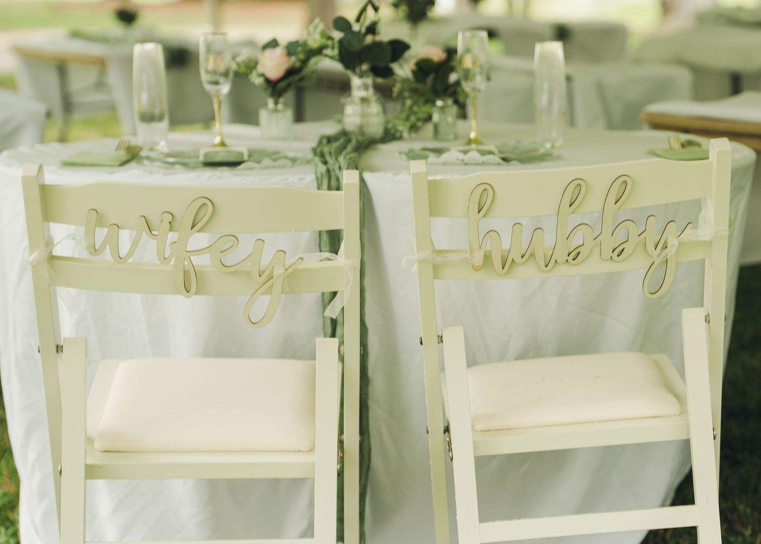 White wedding chairs with gold wifey and hubby signs at an elegant outdoor reception table.