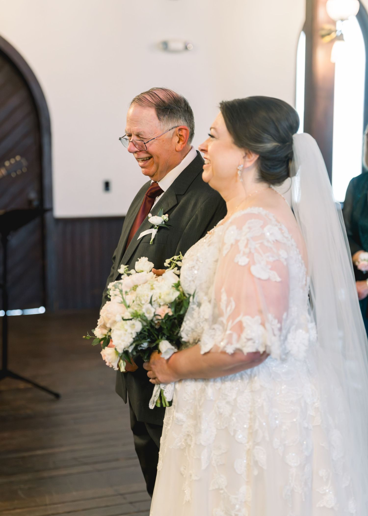 Smiling bride in lace wedding gown holding white bouquet walks down aisle with proud father.
