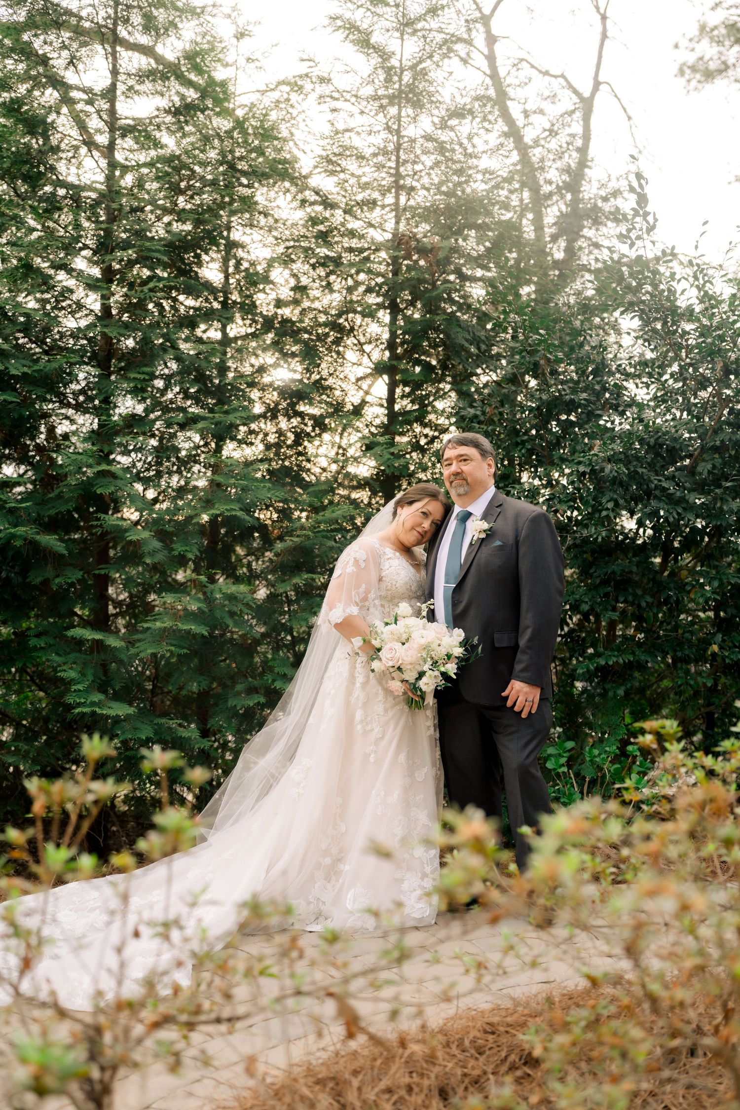 Bride and groom posing together in front of tall evergreen trees with soft natural lighting.