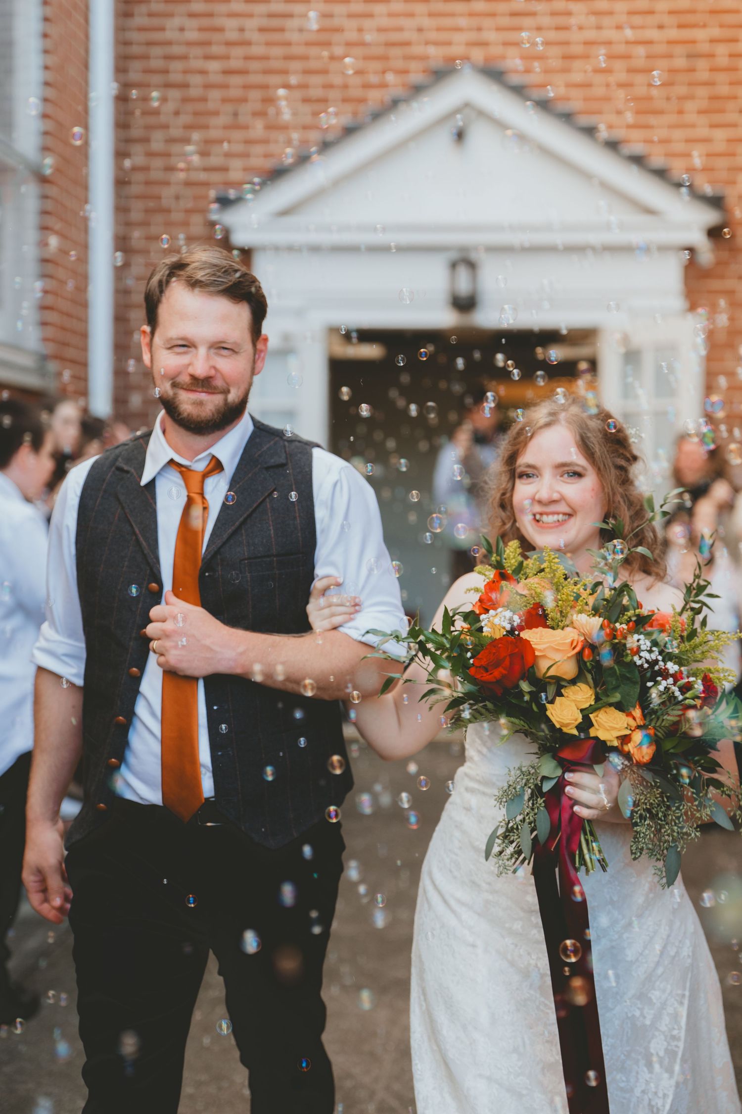 Smiling newlyweds exit a brick church amid a shower of bubbles, bride holding a vibrant autumn bouquet.