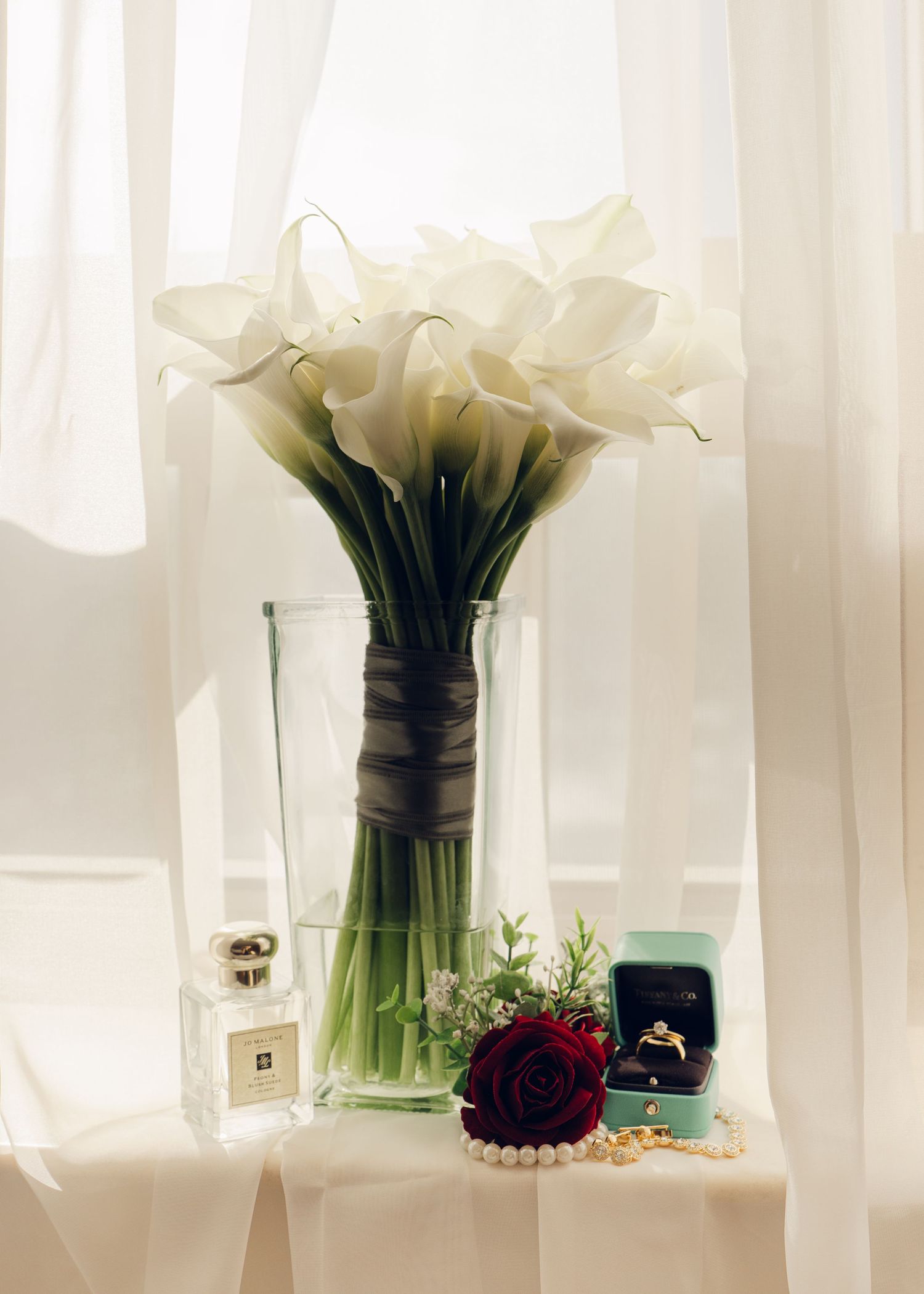 Elegant wedding flat lay featuring white calla lily bouquet, diamond ring, pearl necklace, and red rose.