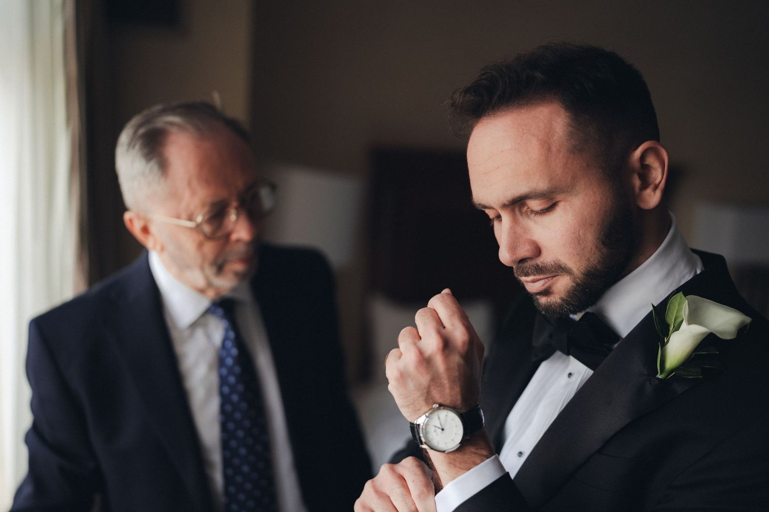 Groom in black tuxedo adjusting watch cufflinks while older man stands in background.