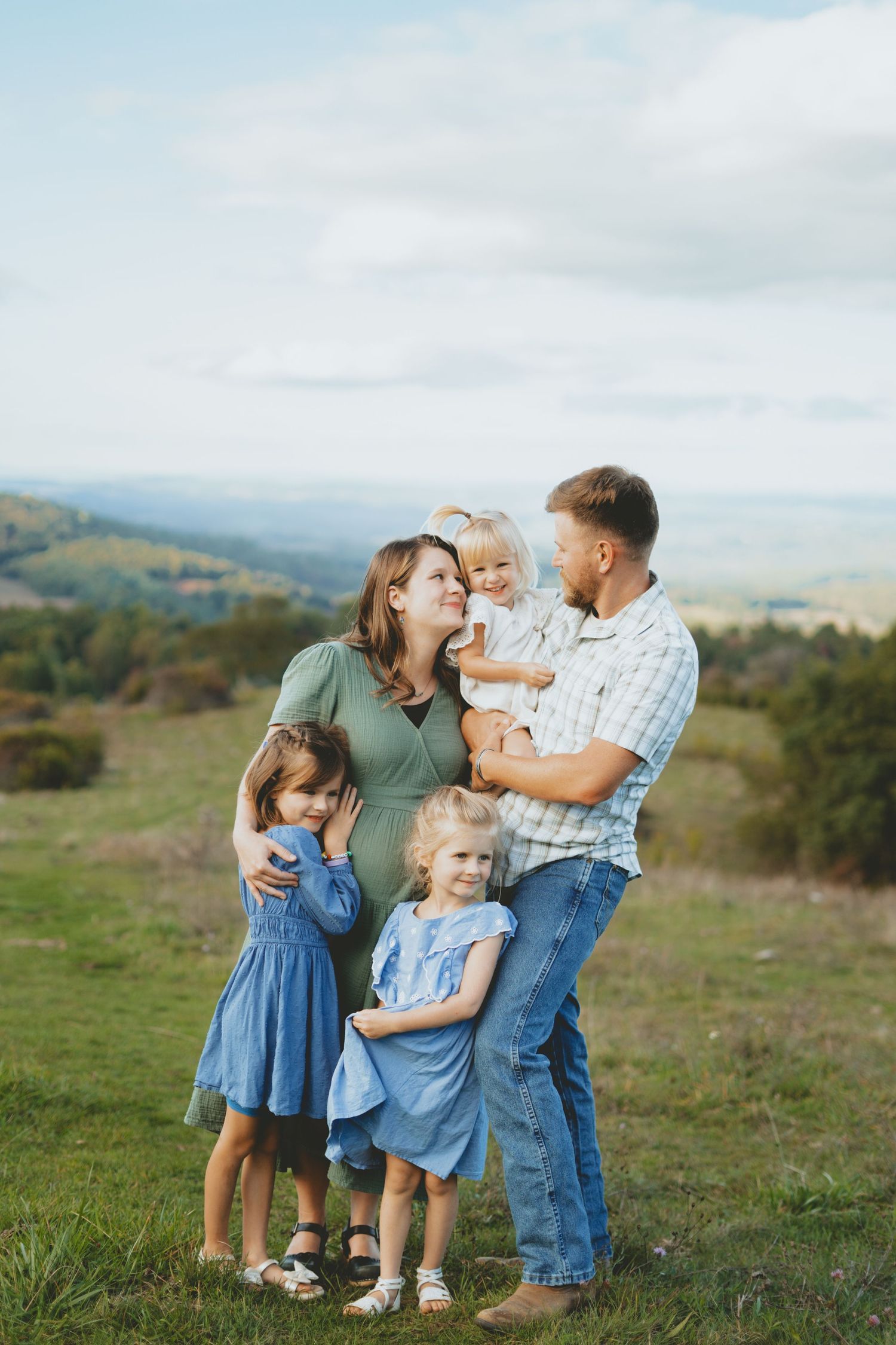 Happy family of five poses outdoors on green hill with scenic valley views, parents holding children in coordinating blue outfits.