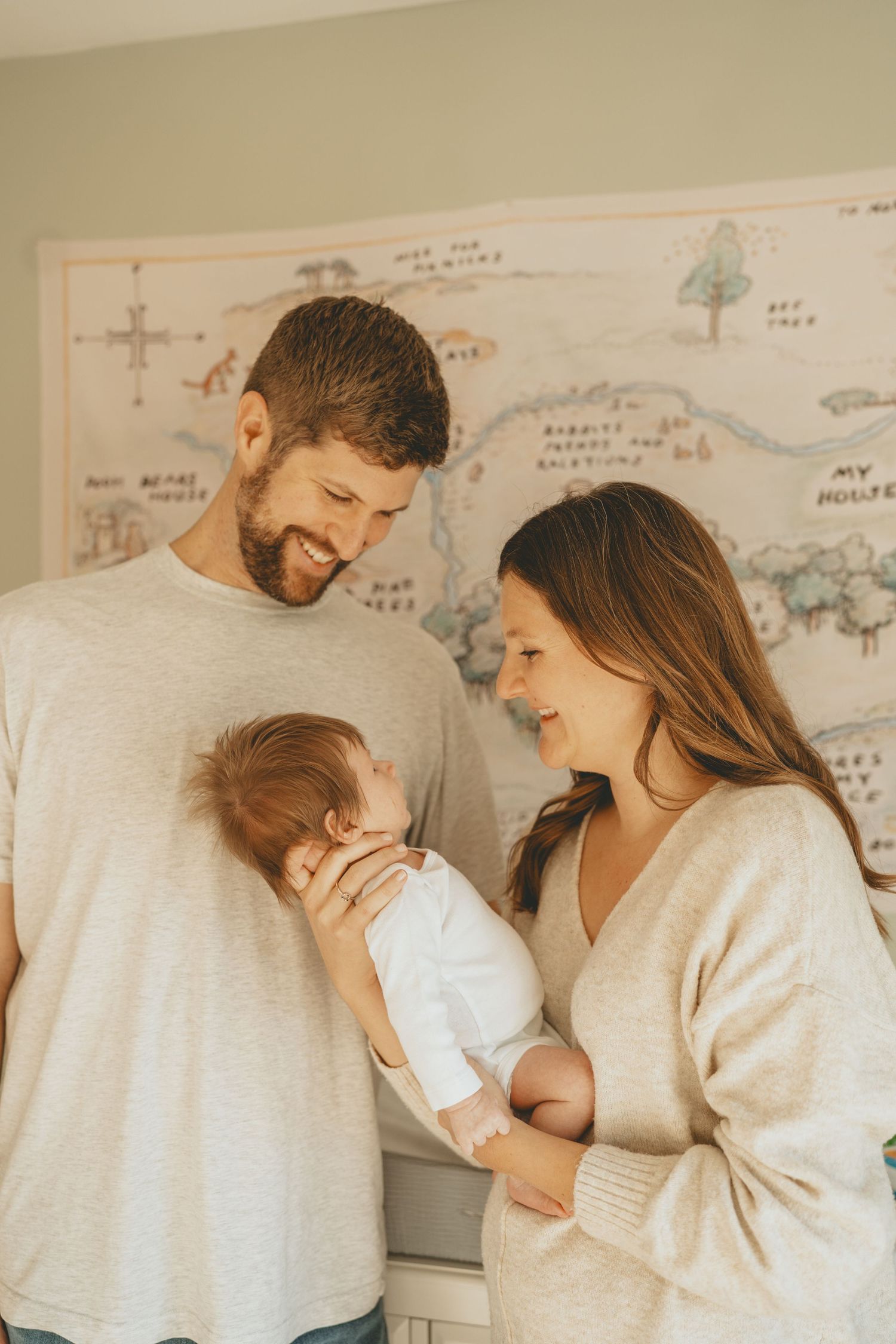 Smiling couple in neutral tones holds their newborn baby in a softly lit nursery with a Winnie the Pooh map.