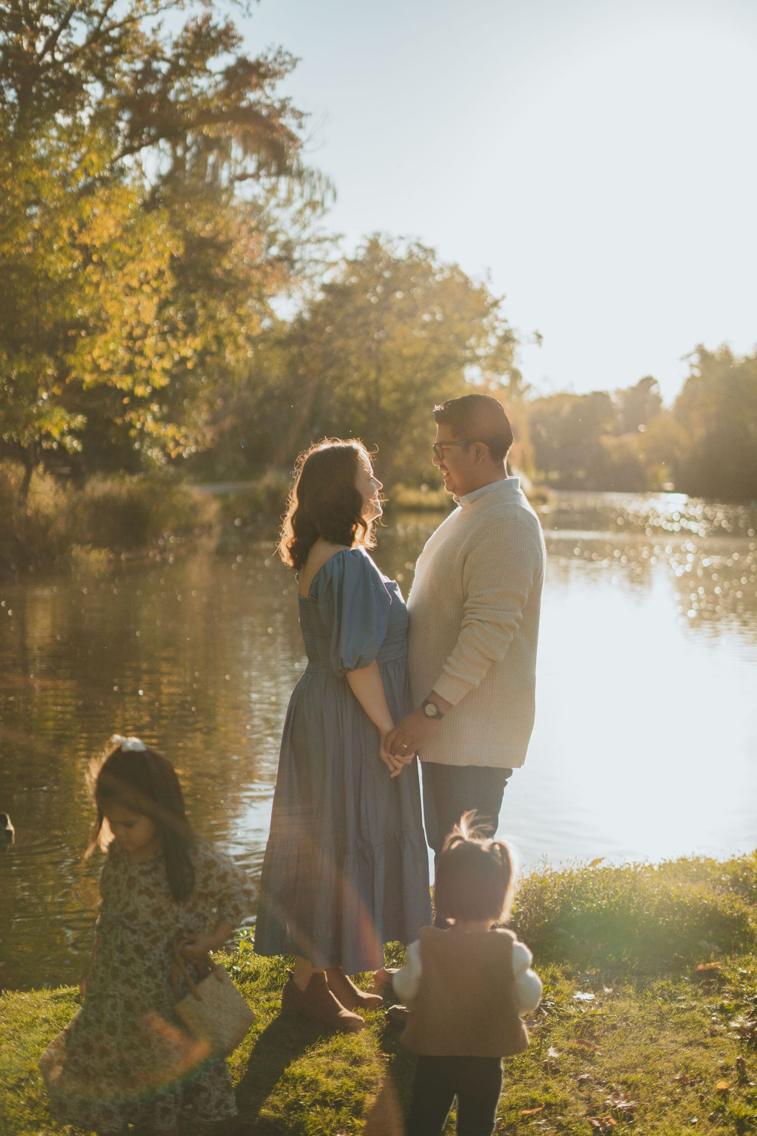 Couple holding hands by a lake while children play in golden sunlight.