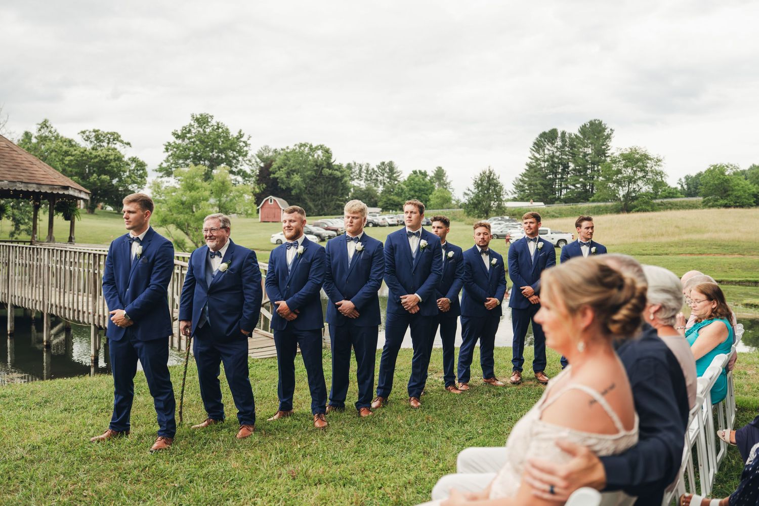 Groomsmen in navy suits standing at an outdoor wedding ceremony on a green lawn.