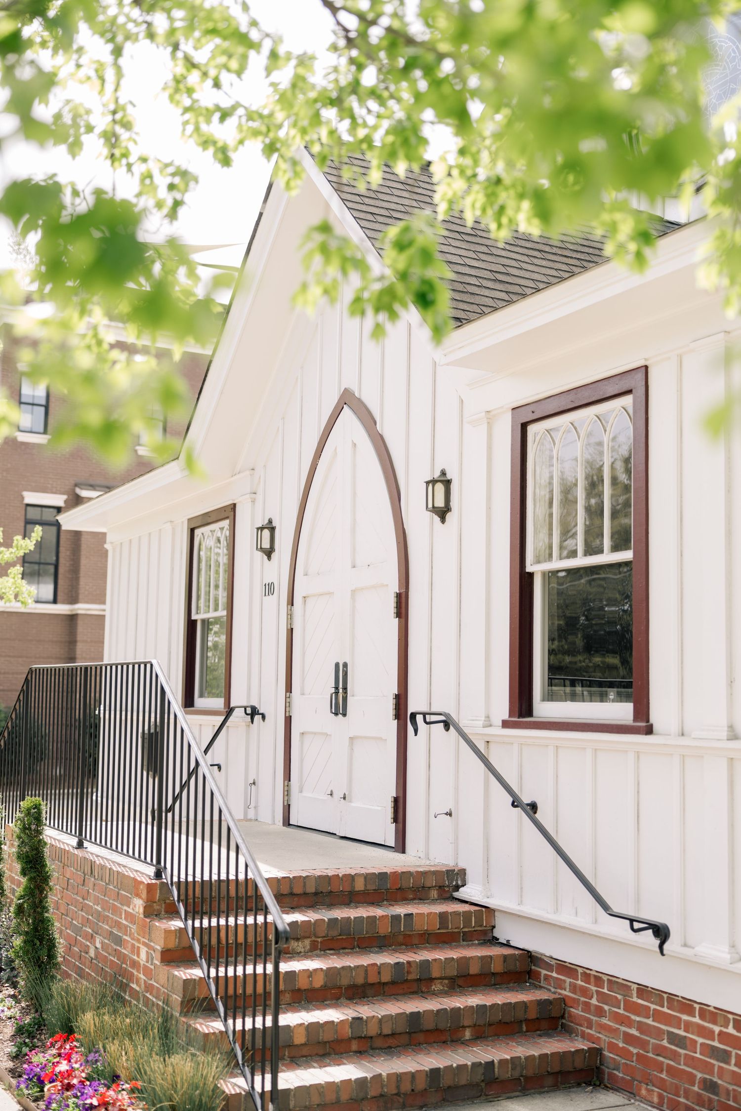 Charming white chapel with gothic arched door, brick steps, and black iron railings framed by green spring foliage.