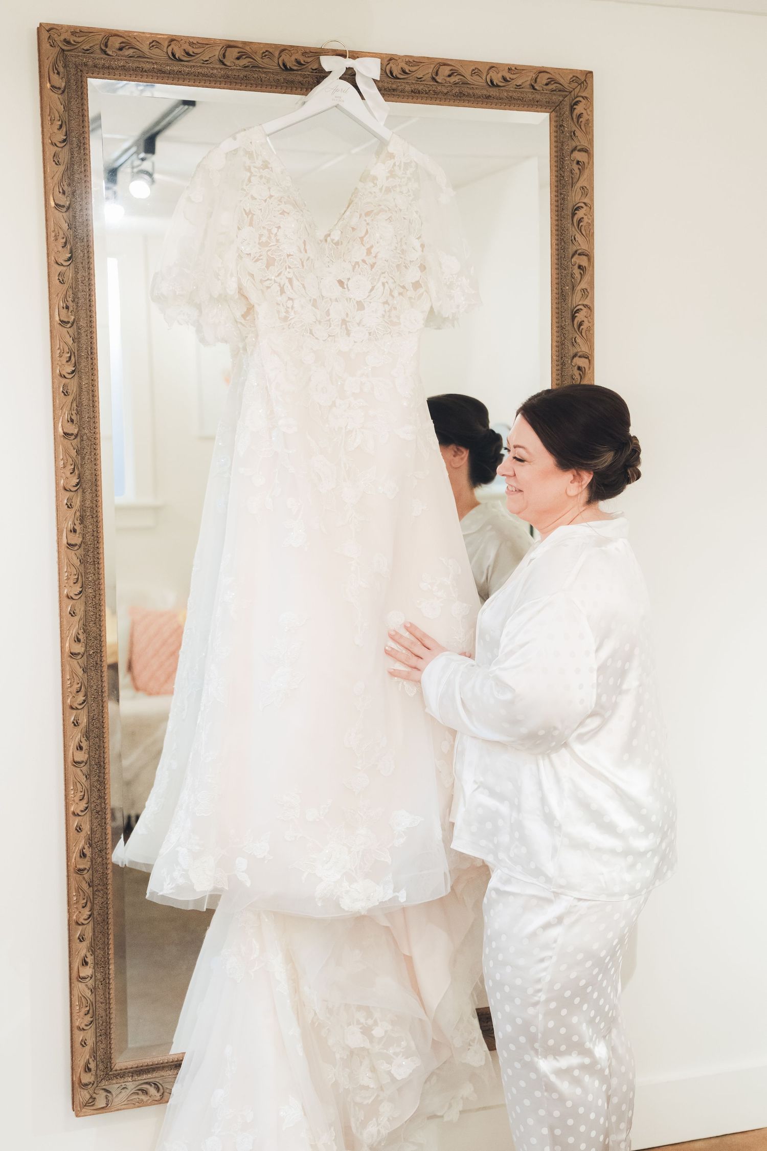 Bride in white pajamas admiring her lace wedding gown hanging in an ornate gold mirror.