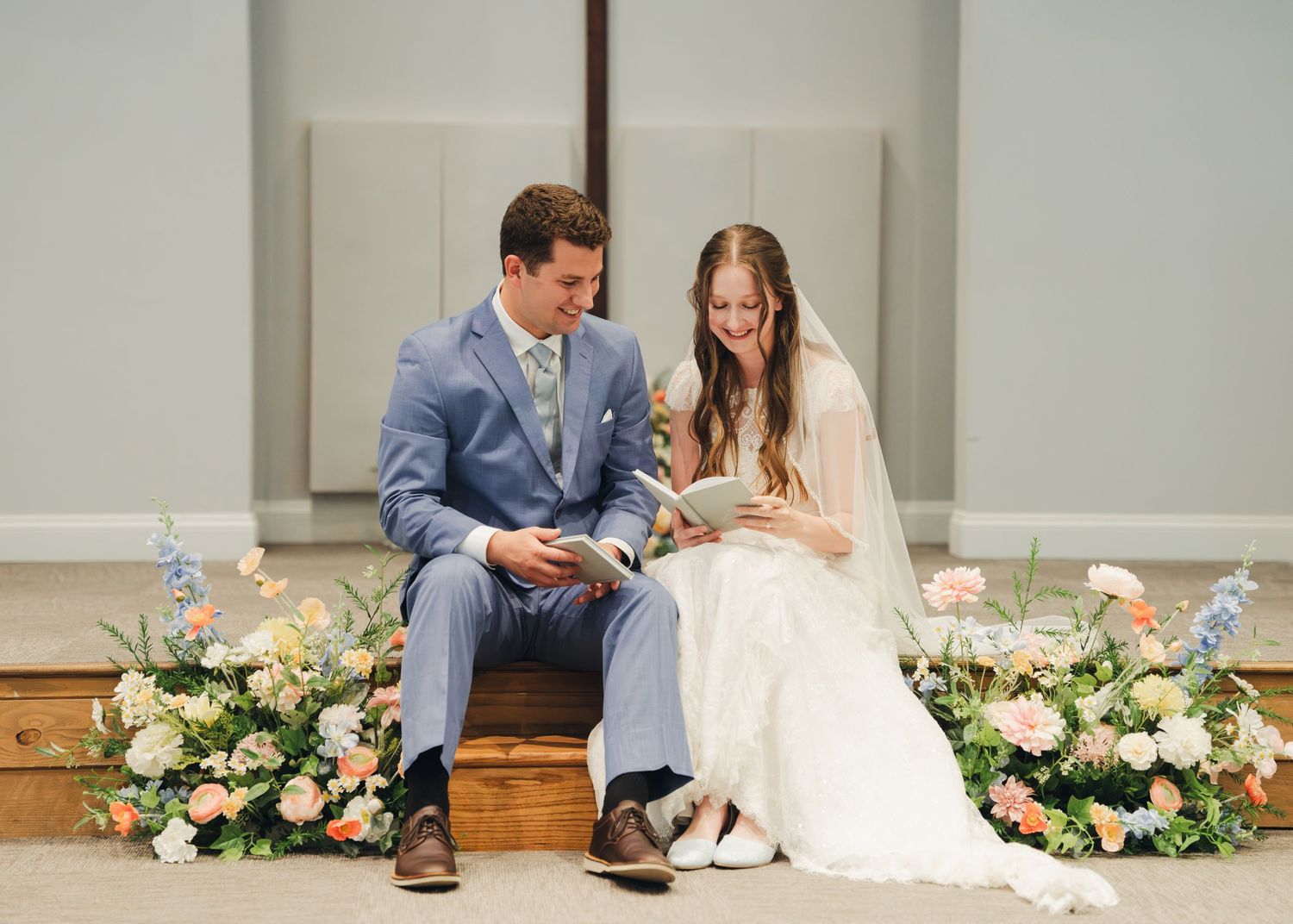 Bride and groom reading vows together, seated on wooden steps surrounded by pastel floral arrangements in a bright chapel.