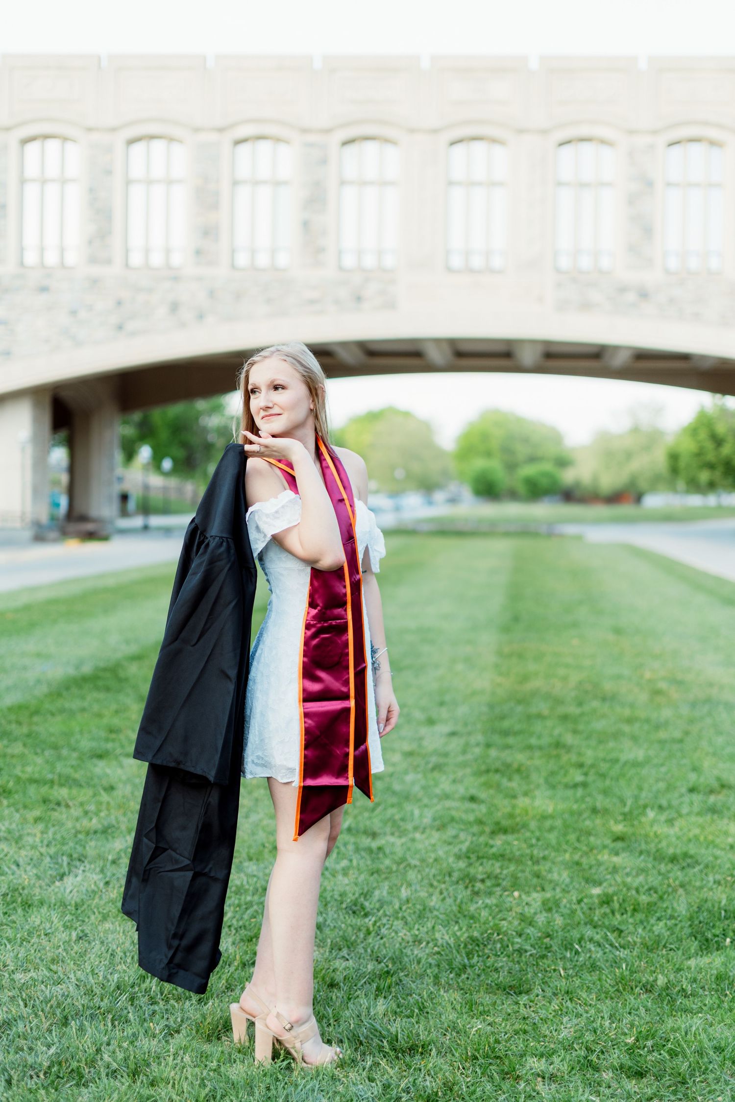 Blonde graduate in white dress carries black gown on green campus lawn at Virginia Tech.
