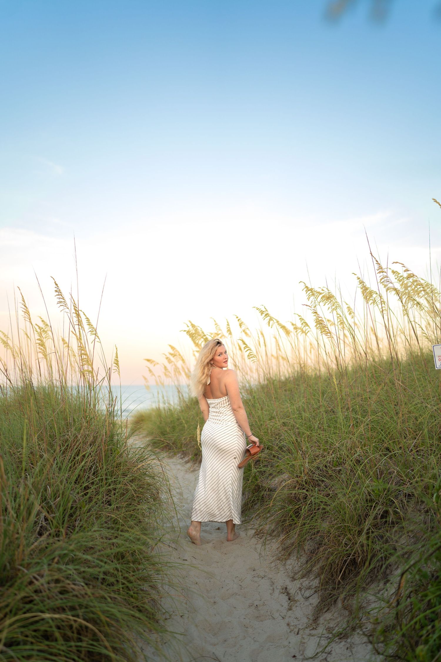 Blonde woman in white polka dot dress on sandy beach path surrounded by sea oats at sunset.