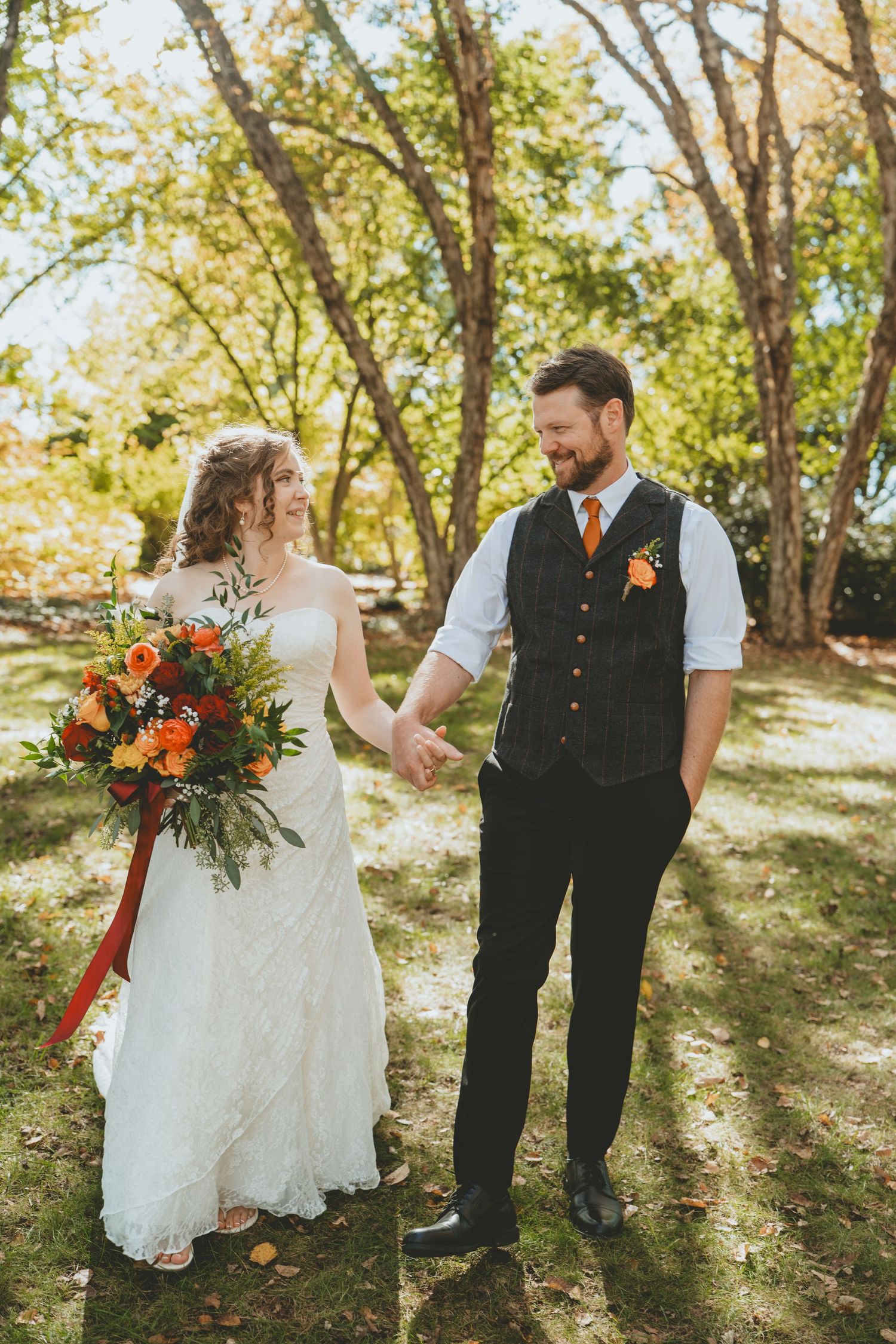 Bride and groom holding hands and smiling at each other in a sunlit autumn garden setting.