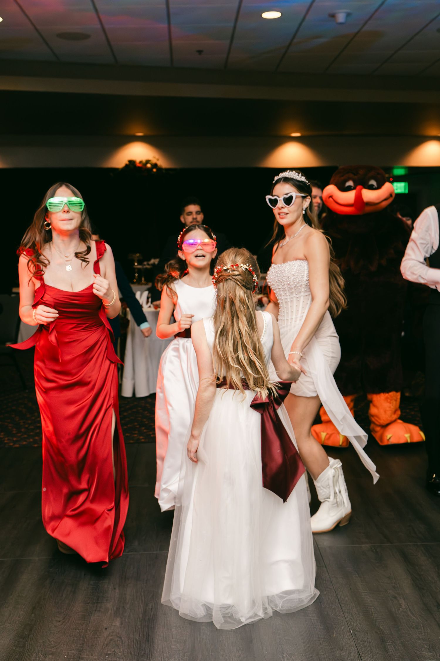 Bride in white mini dress and heart sunglasses dances with flower girl and bridesmaid in red gown at wedding reception.