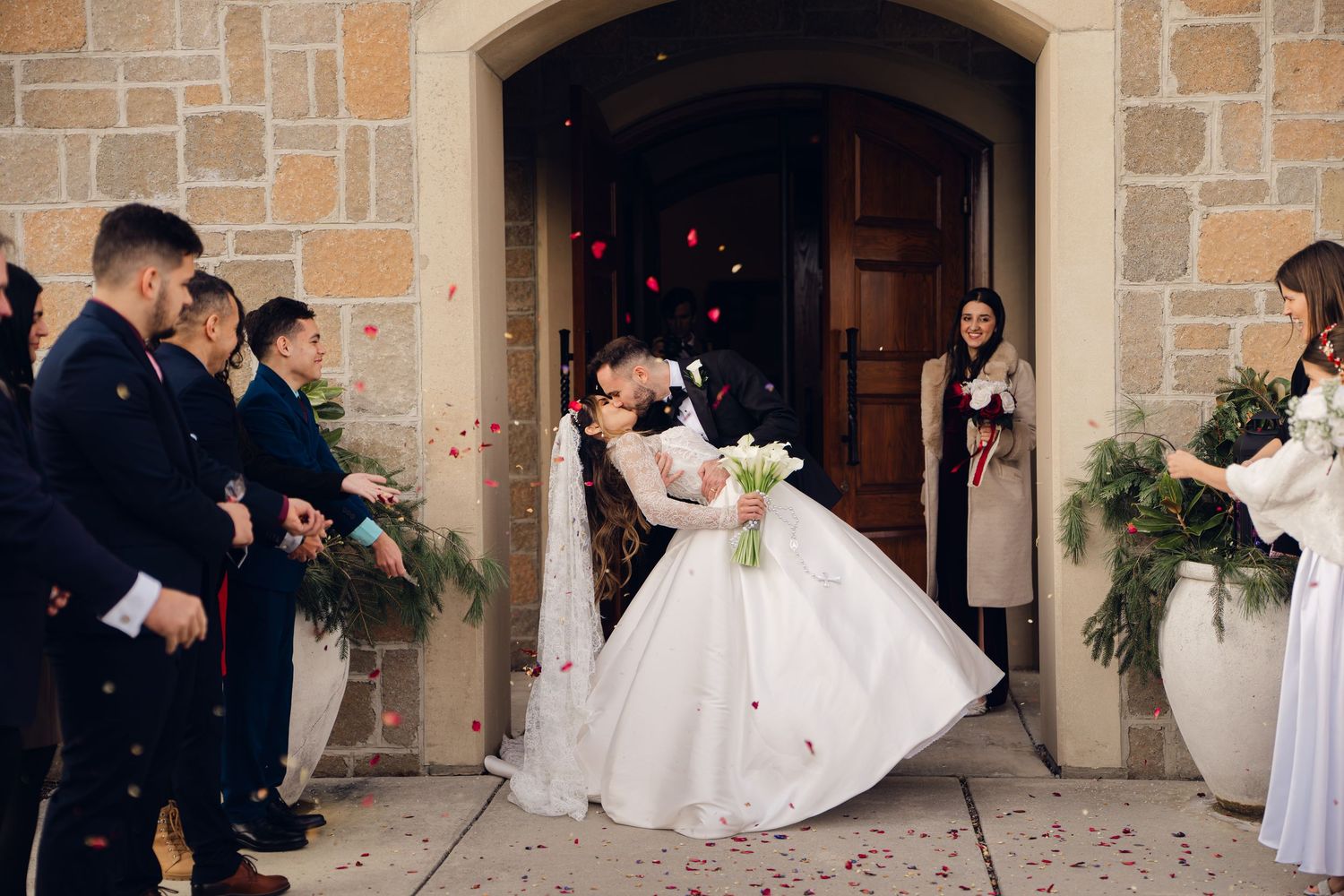 Groom dips bride for romantic kiss outside stone church as guests toss rose petals.