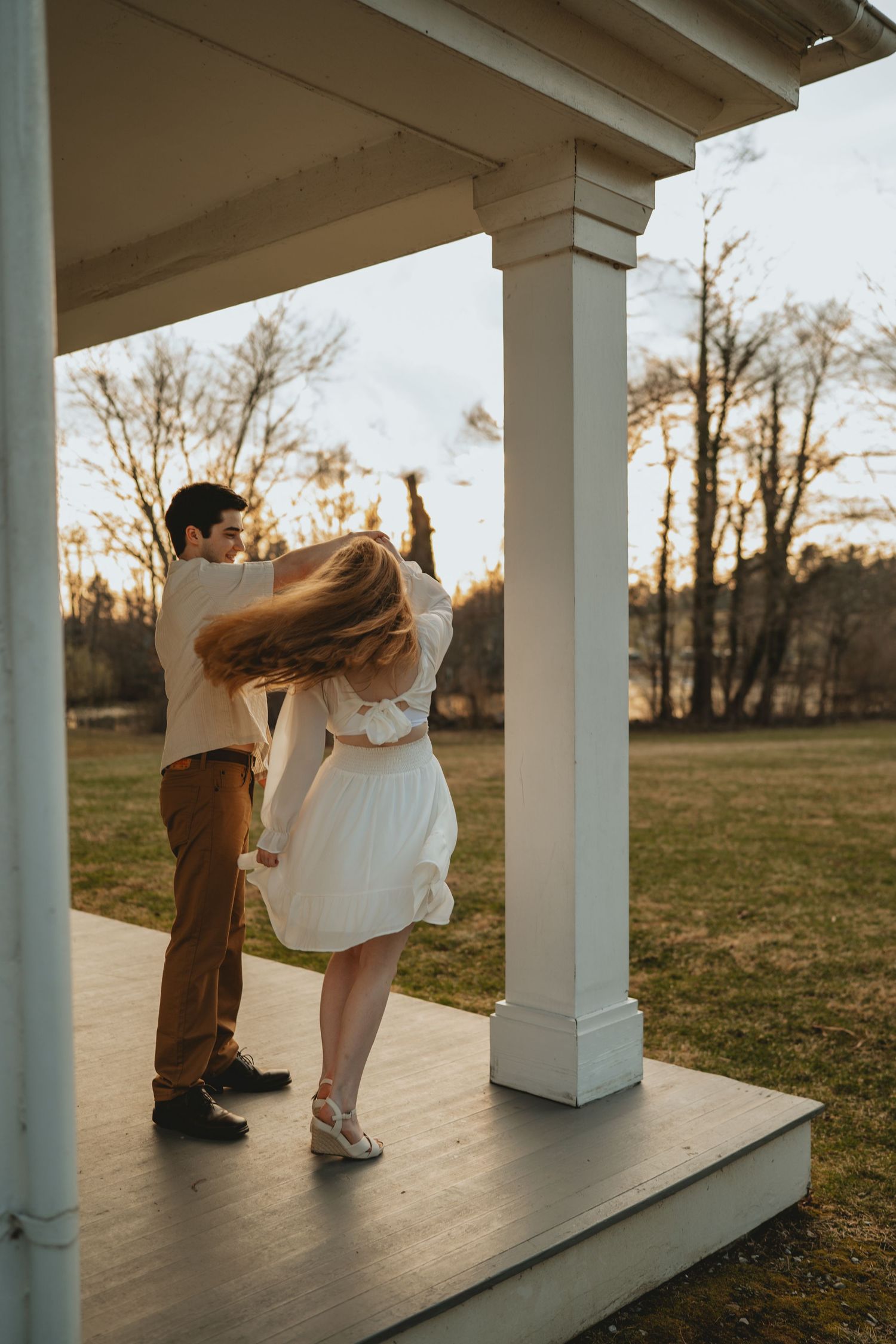 Couple dancing and twirling on a white porch at sunset with golden light and bare trees.