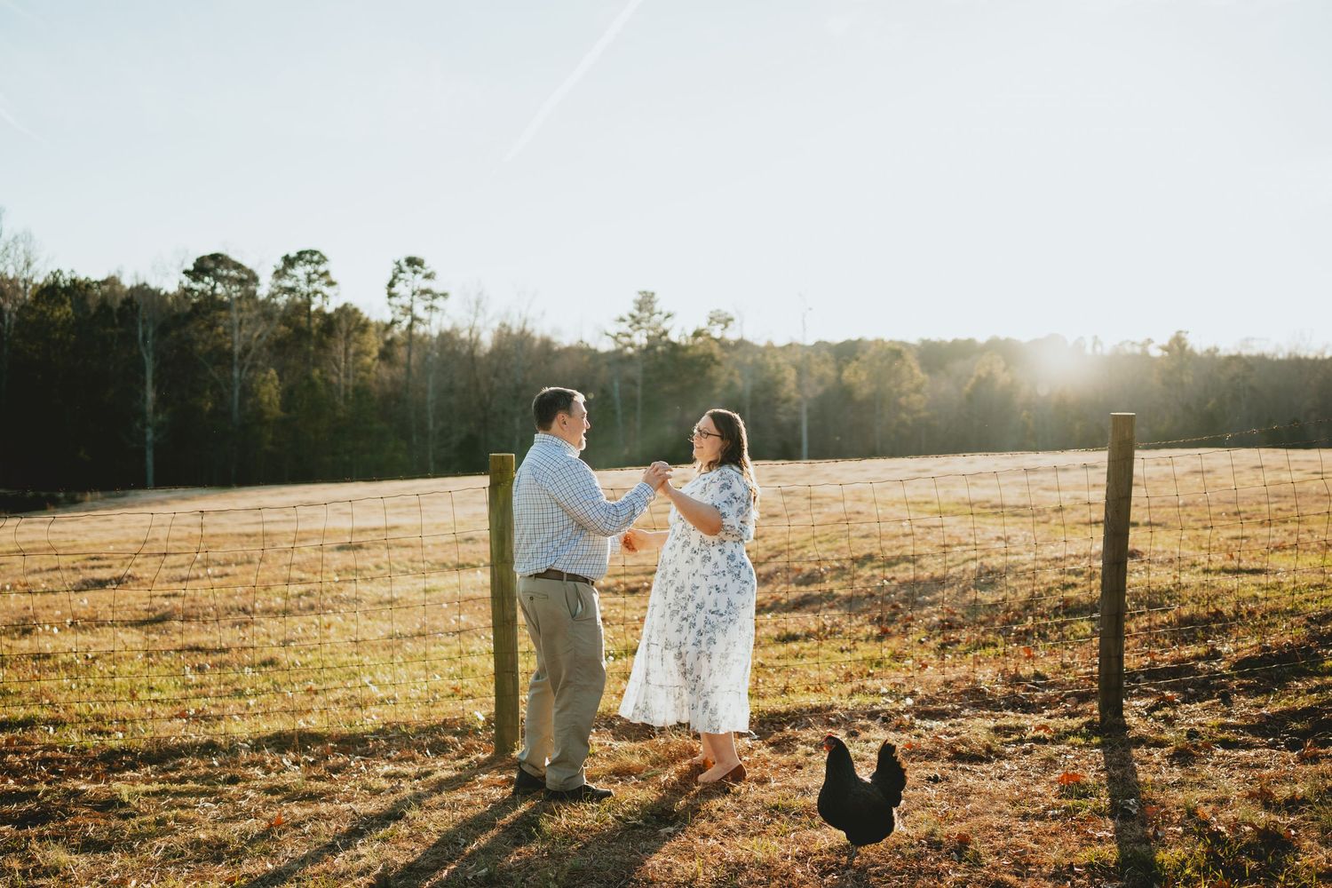 Joyful couple dancing together in a sun-drenched farm field with a rooster nearby.