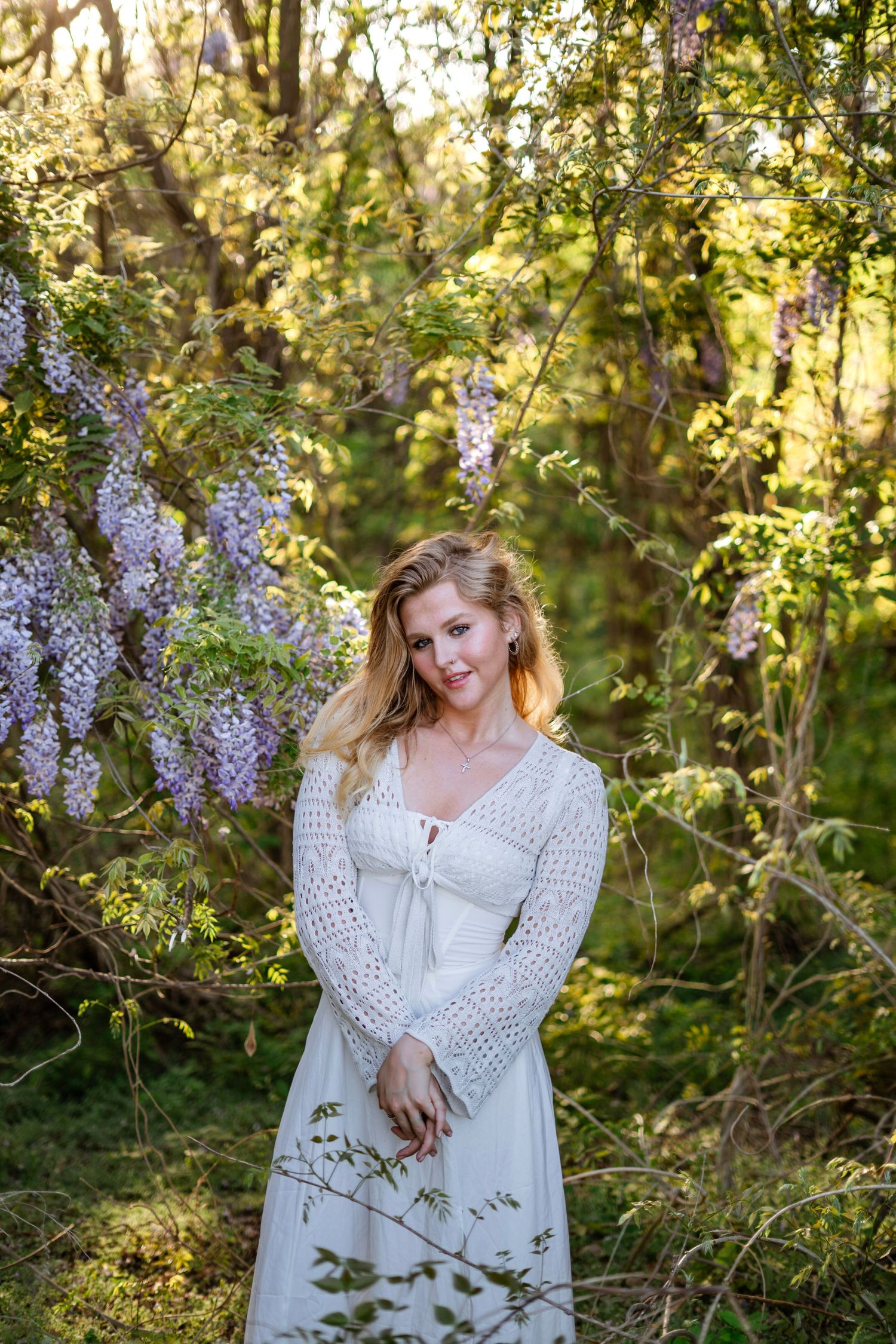 Blonde woman in white lace dress standing among purple wisteria blooms in sunlit garden.