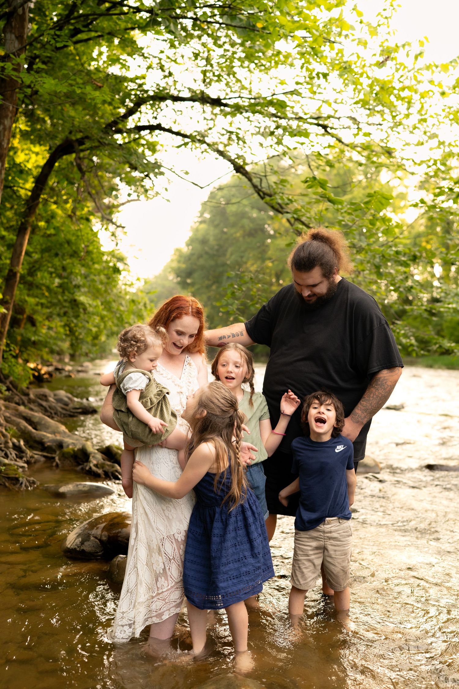 Family of six laughing and playing together while standing in a shallow creek surrounded by trees.