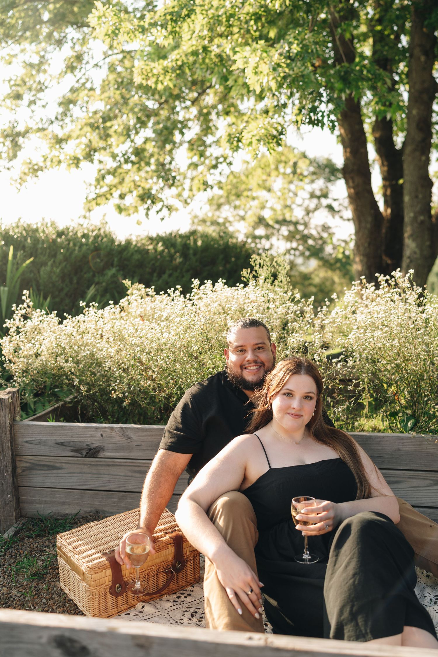Couple relaxing at a picnic with wine and a wicker basket surrounded by wildflowers in golden hour light.