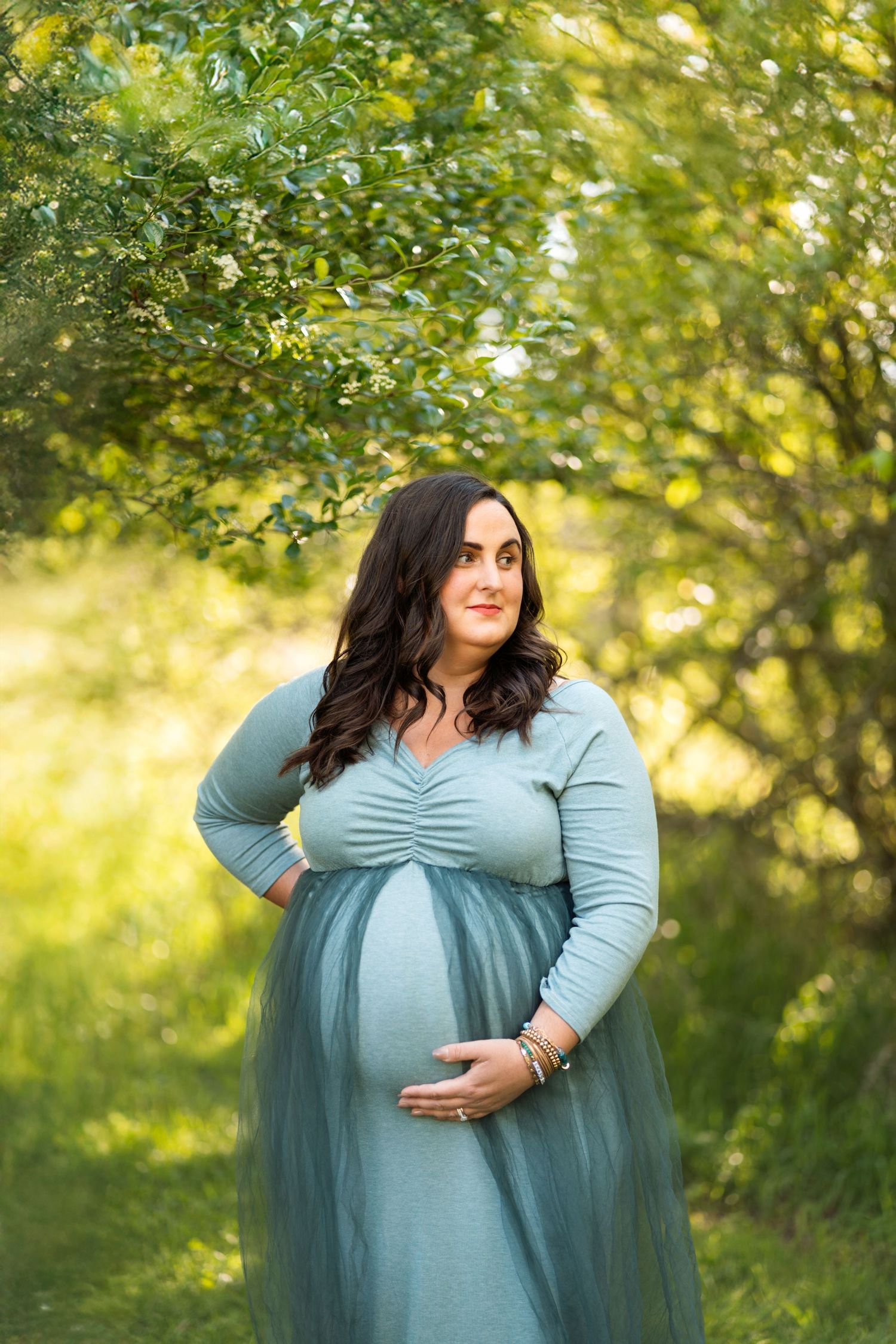 A pregnant woman in a sage green gown cradles her baby bump in a lush green outdoor setting.