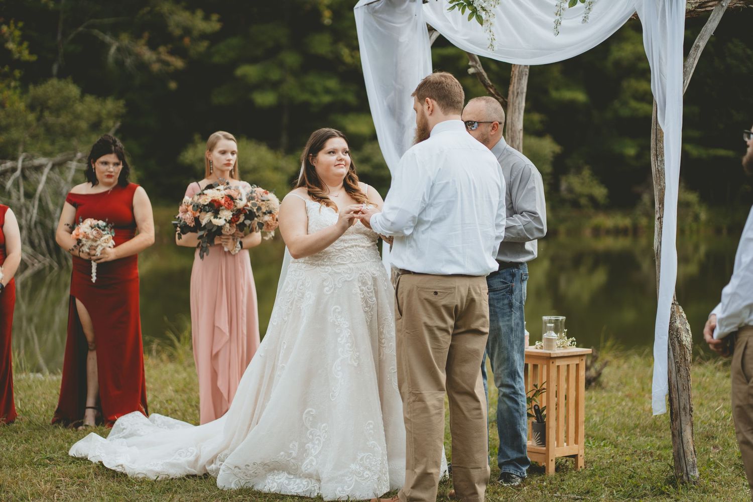 Bride exchanges rings with groom at outdoor wedding altar surrounded by bridesmaids in blush and red gowns.