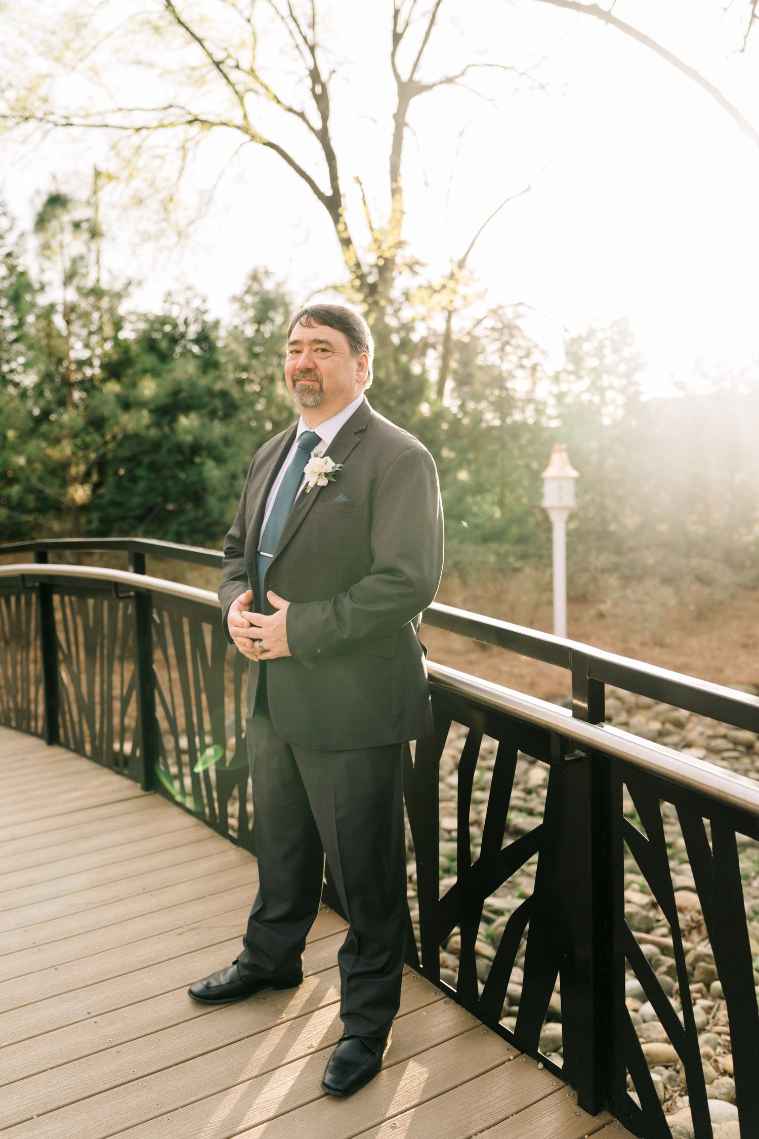 Groom in dark suit with boutonniere stands on wooden bridge in sunlit garden.
